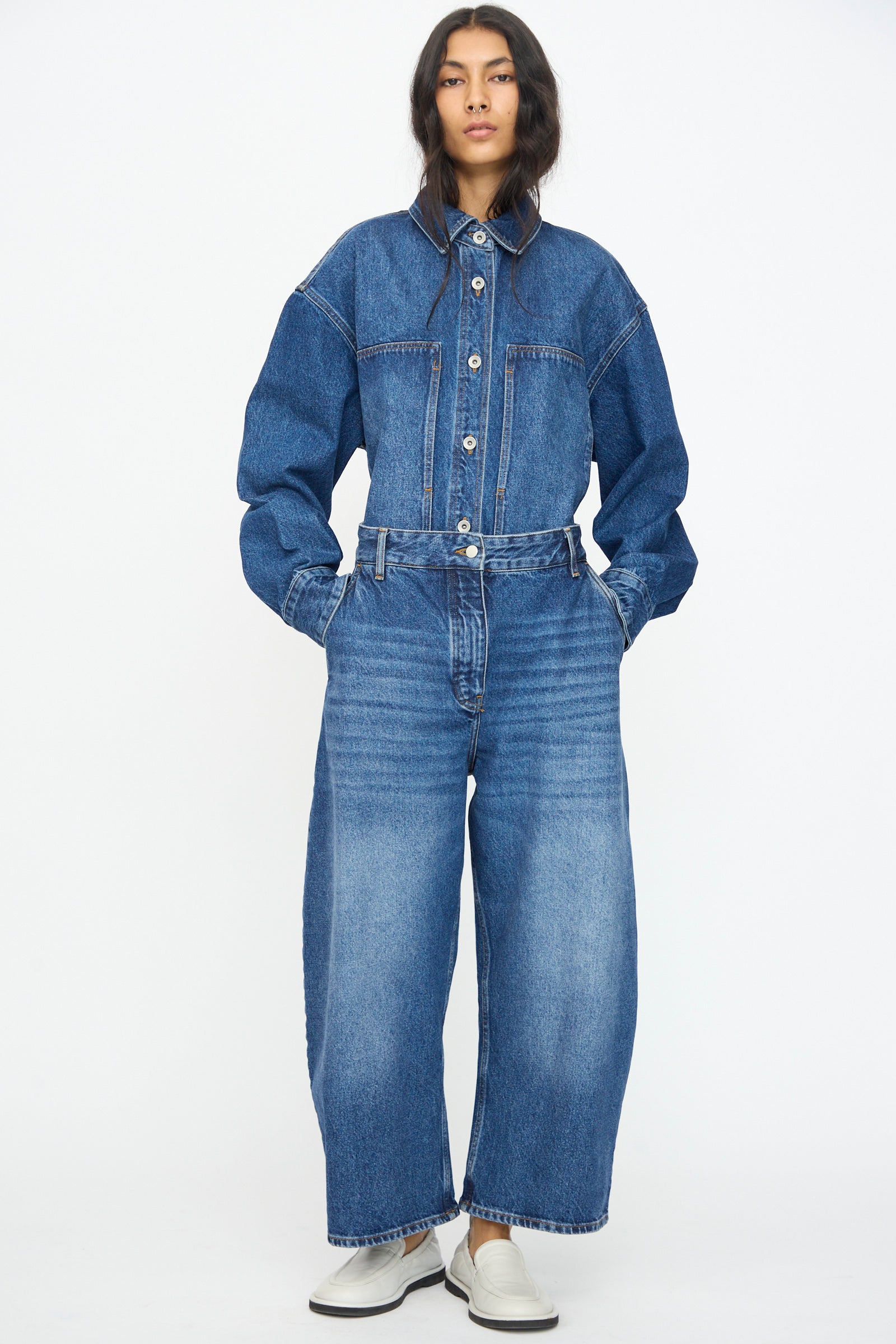 A person stands against a white background wearing wide leg Studio Nicholson Chalco Jeans in Worn Wash Indigo, paired with an oversized washed denim shirt and white shoes, hands in pockets.