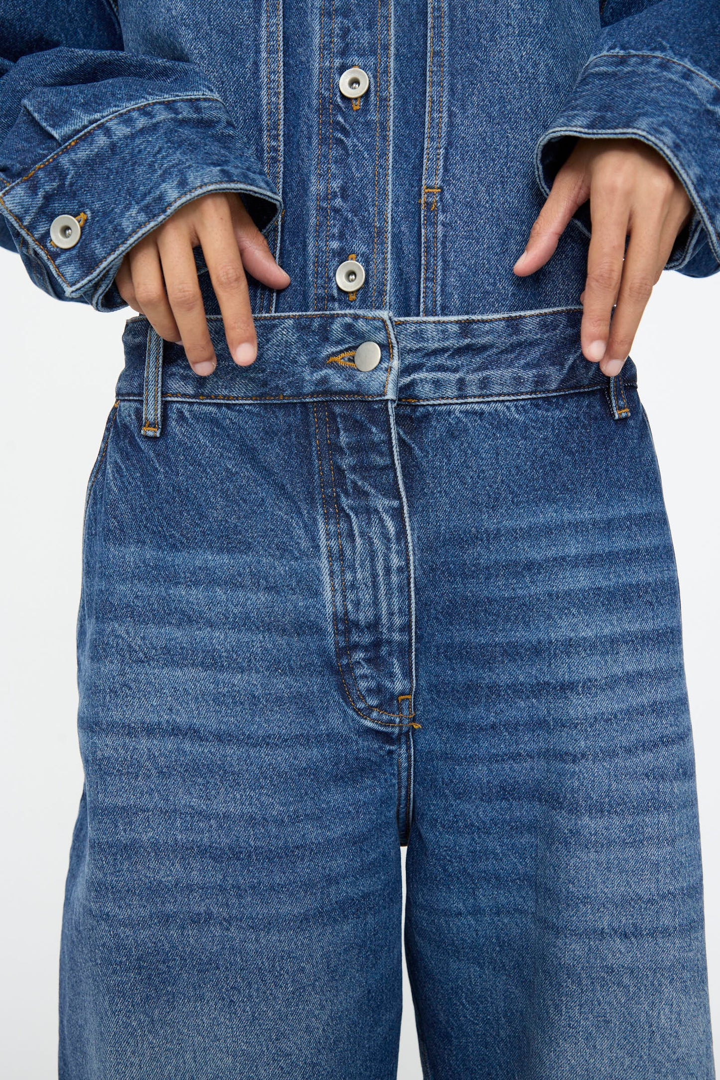 Model shown from the chest down wearing Studio Nicholson's Chalco Jean in Worn Wash Indigo, paired with a matching jacket and holding the jeans' waistband with both hands.