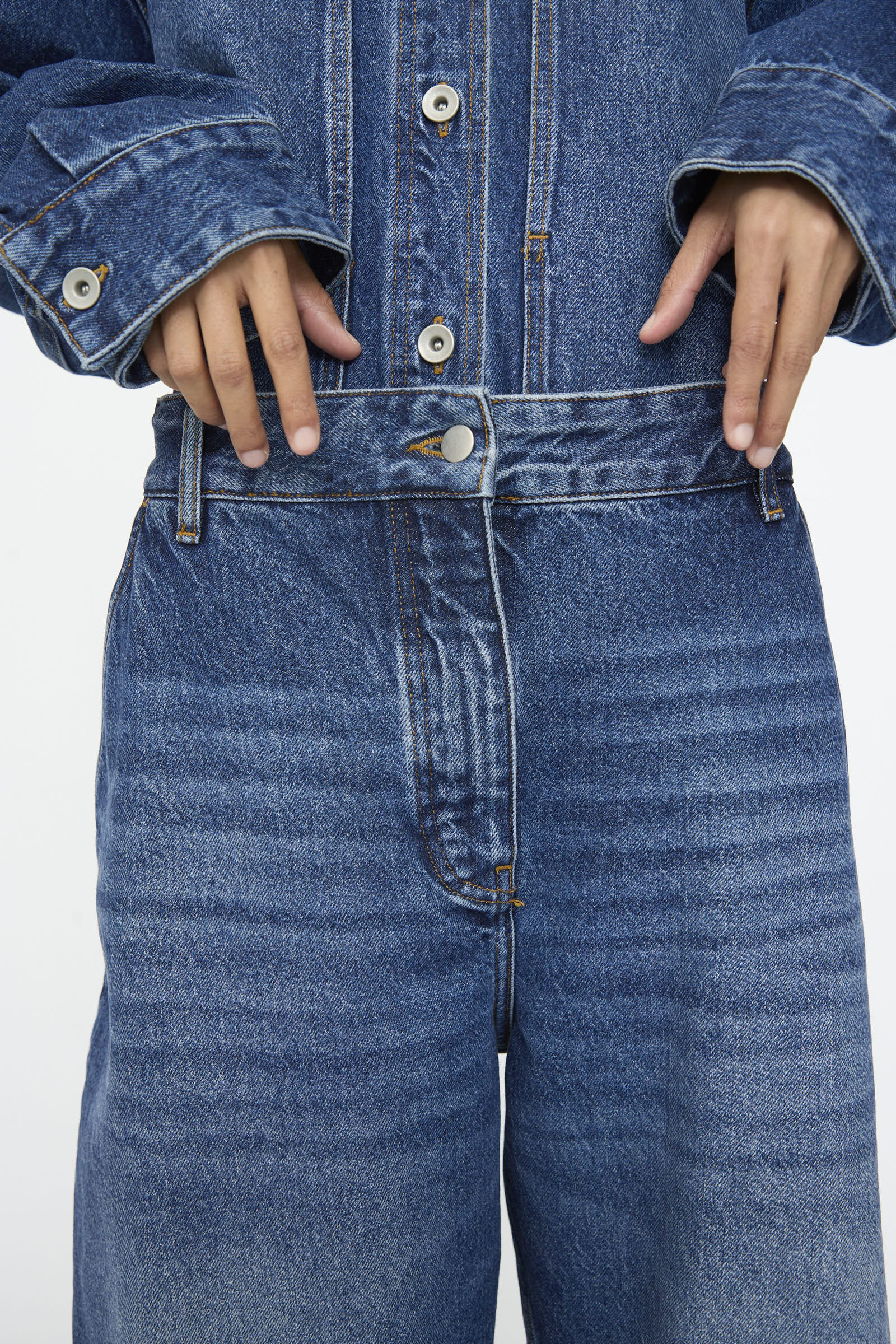 Model shown from the chest down wearing Studio Nicholson's Chalco Jean in Worn Wash Indigo, paired with a matching jacket and holding the jeans' waistband with both hands.