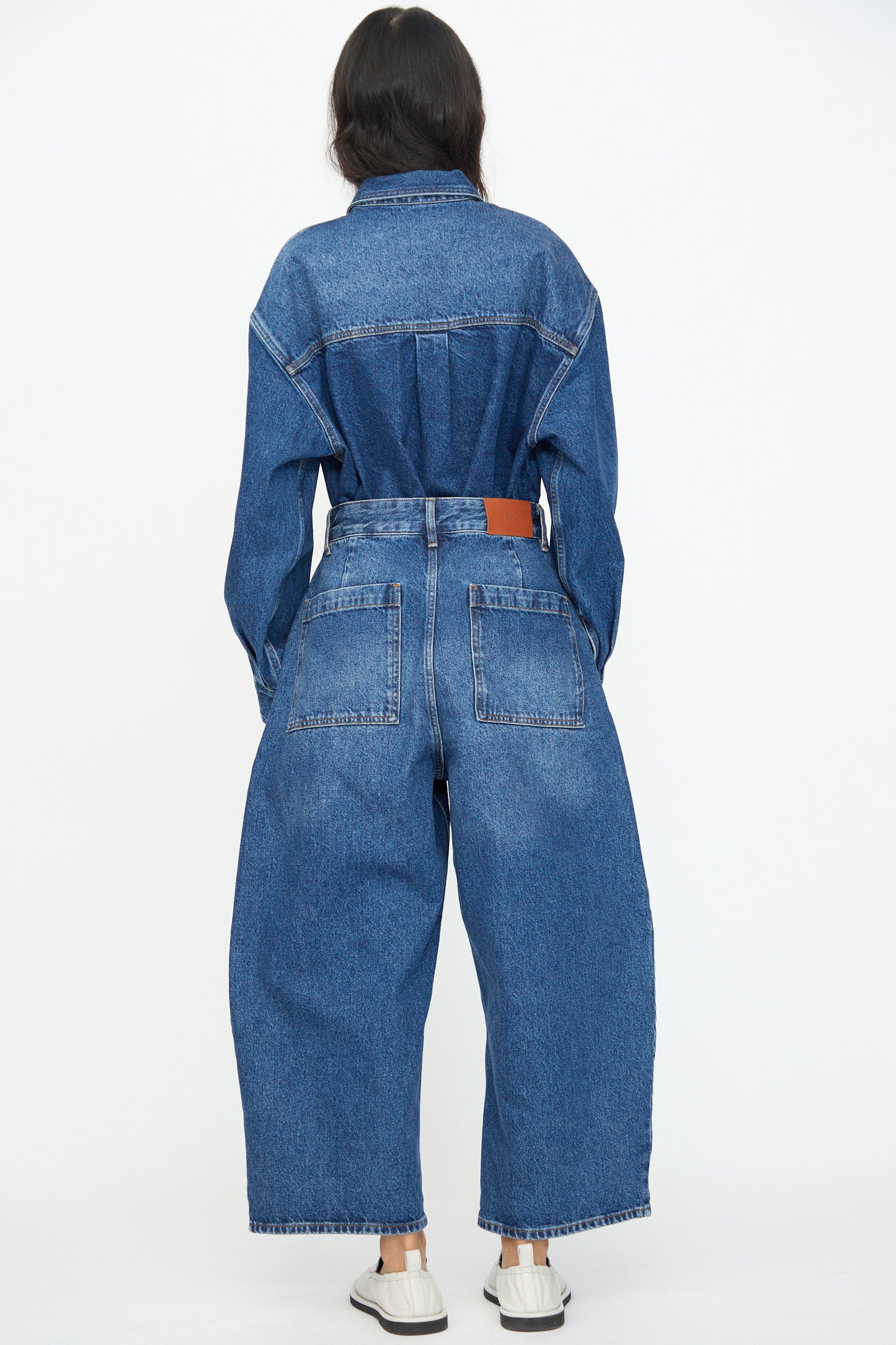 A person faces away, wearing Studio Nicholson's Chalco Jean in Worn Wash Indigo—an oversized fit denim jumpsuit with wide legs—paired with white slip-on shoes, set against a plain white background.