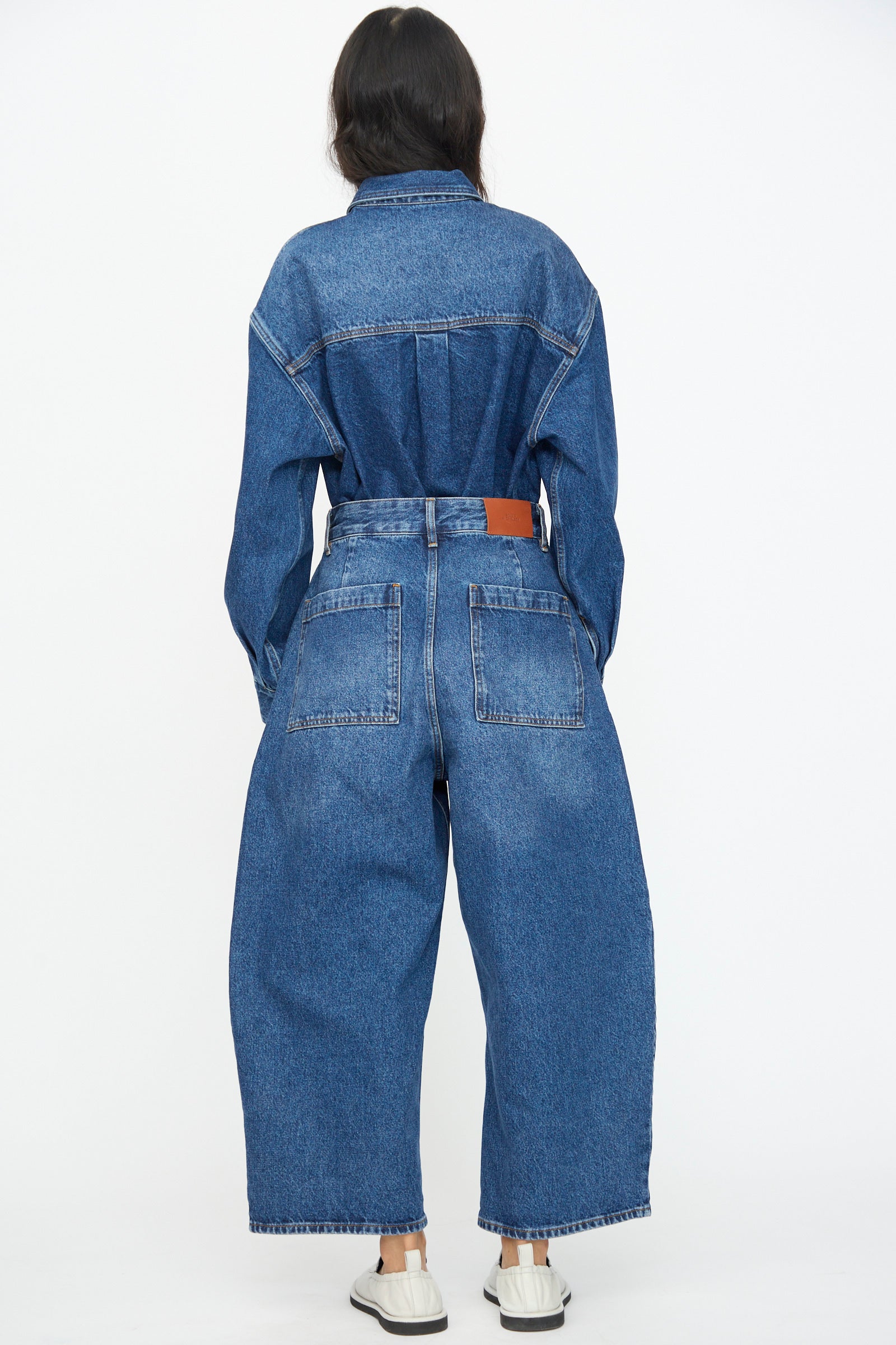 A person faces away, wearing Studio Nicholson's Chalco Jean in Worn Wash Indigo—an oversized fit denim jumpsuit with wide legs—paired with white slip-on shoes, set against a plain white background.