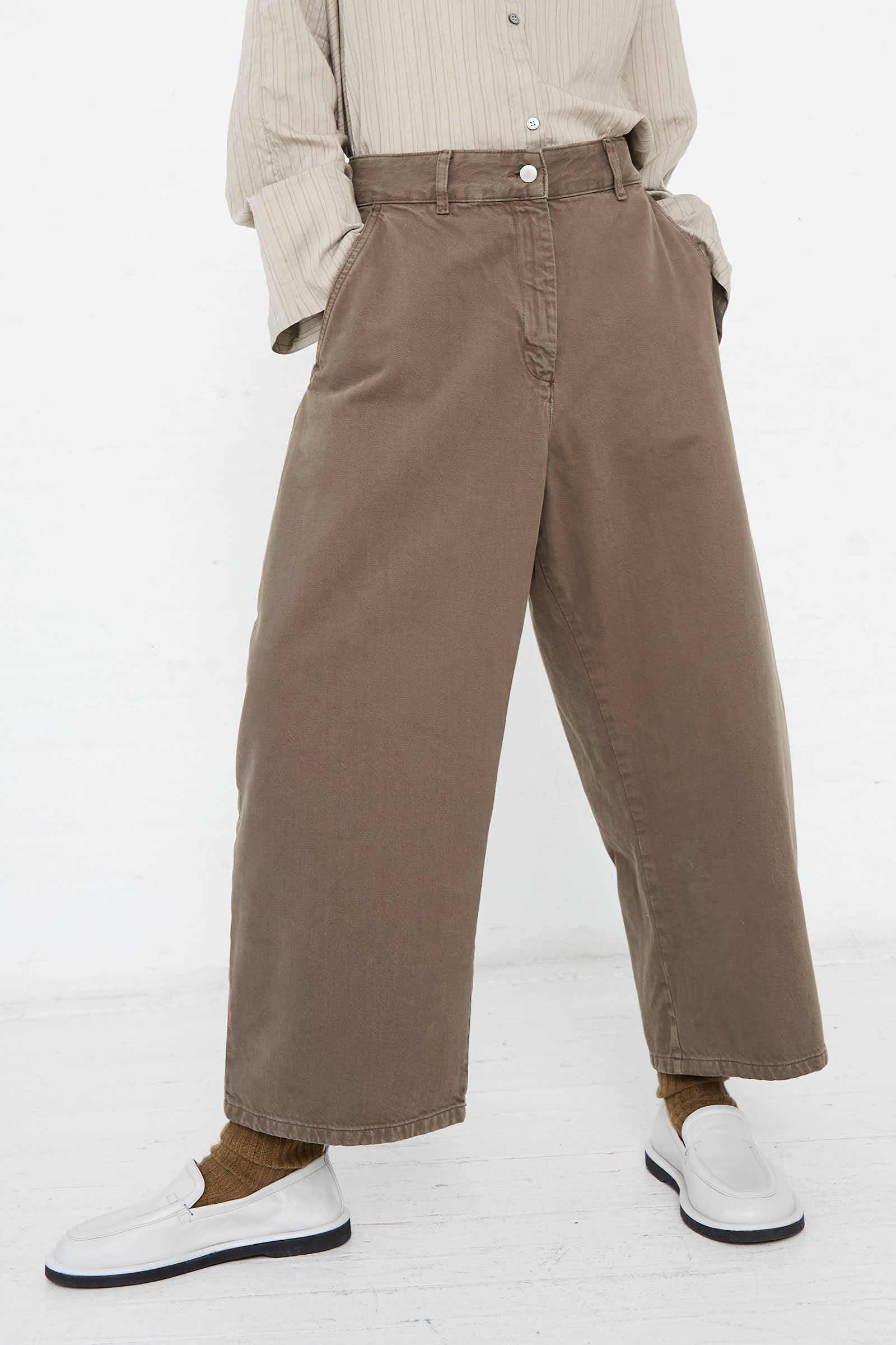 A person is wearing a beige long-sleeve shirt tucked into the Studio Nicholson Chalco Wide Crop Pant in Fossil, made from recycled brown denim material, with hands in pockets. Paired with white shoes and brown socks, the relaxed fit ensemble stands out against a white background.