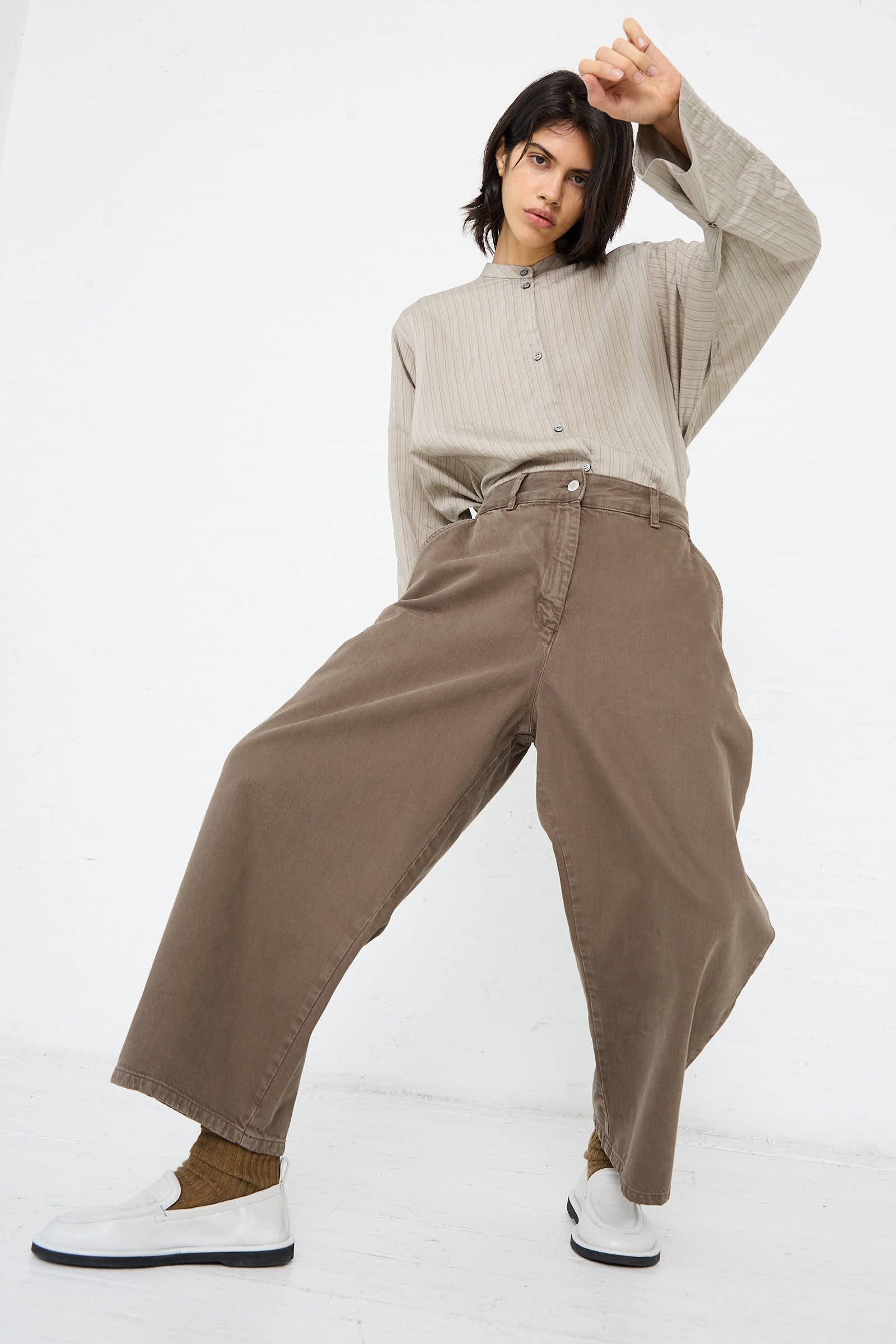 Person wearing an oversized beige top and the Chalco Wide Crop Pant in Fossil from Studio Nicholson, made from recycled material, with one arm raised and looking at the camera, standing against a plain white background.