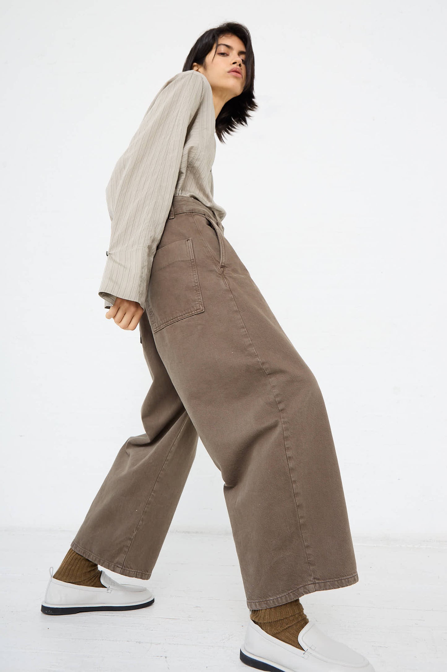 Person wearing a beige long-sleeve top, Studio Nicholson's Chalco Wide Crop Pant in Fossil made from recycled material, brown socks, and white slip-on shoes, tilting body slightly back and leaning to the right. Neutral background.