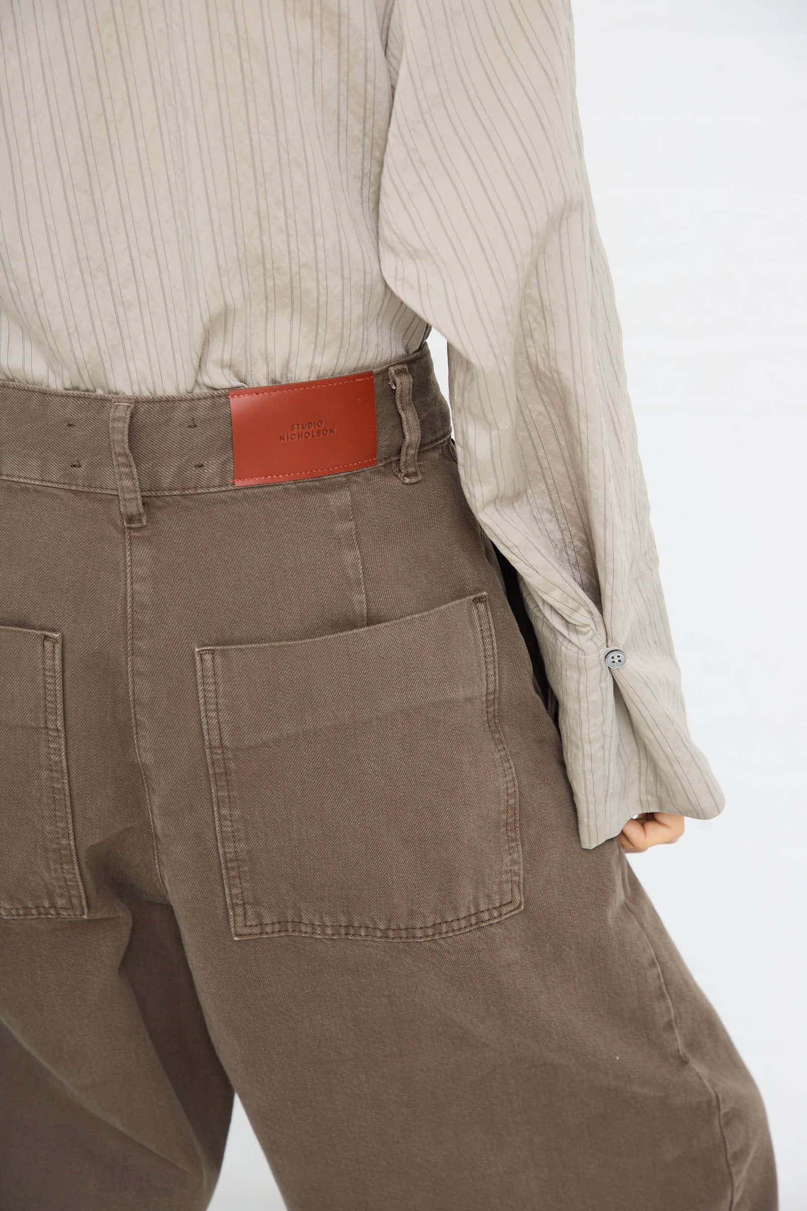 Back view of a person wearing a beige pinstripe shirt and the Studio Nicholson Chalco Wide Crop Pant in Fossil with oversized pockets.