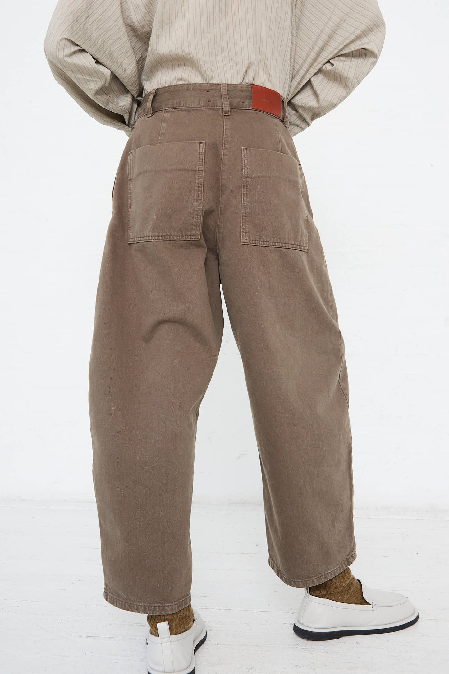 A person wearing Studio Nicholson's Chalco Wide Crop Pant in Fossil with a relaxed fit, a beige striped shirt, and white shoes stands with their back facing the camera.