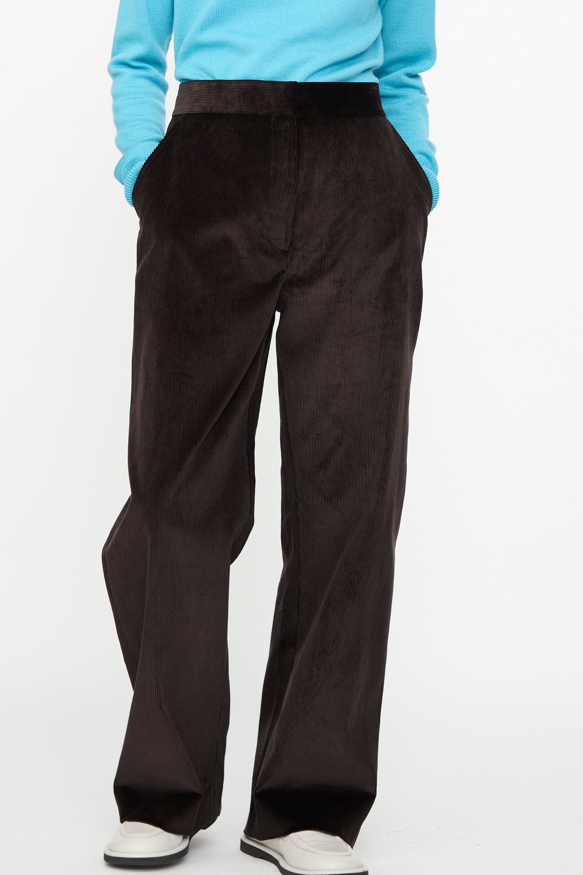A person stands with hands in pockets against a plain background, wearing Studio Nicholson’s Cotton Corduroy Grange Curved Pant in Espresso—featuring a relaxed, curved leg silhouette—paired with a light blue long-sleeve cotton sweater and white shoes.