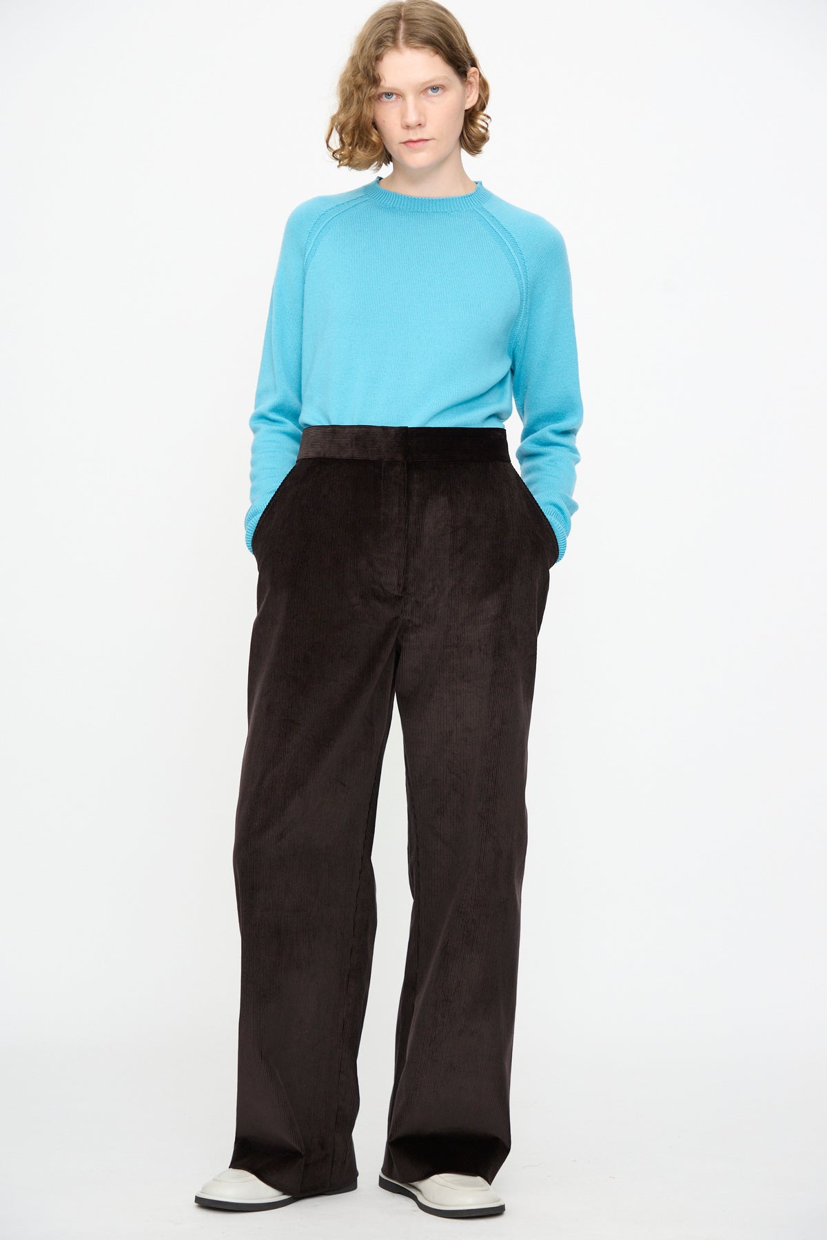 A person wearing a light blue sweater, Studio Nicholson’s Cotton Corduroy Grange Curved Pant in Espresso—a high-waisted, wide-leg style with a curved leg silhouette—and white shoes stands against a plain white background.
