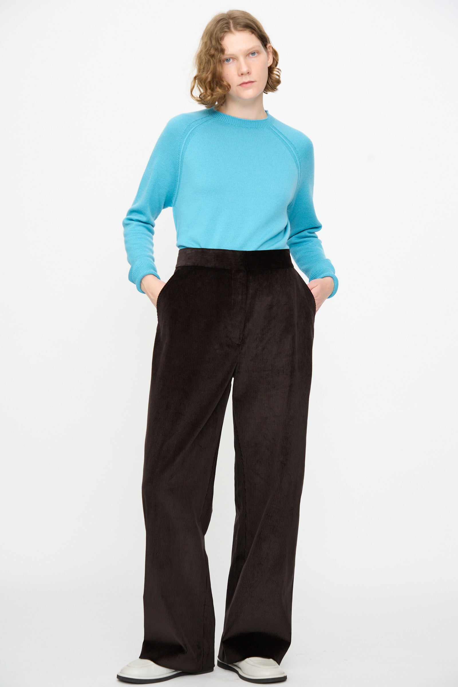 A person stands against a white background, hands casually tucked in their pockets, wearing a light blue sweater and the Studio Nicholson Cotton Corduroy Grange Curved Pant in Espresso, paired with white cotton shoes.