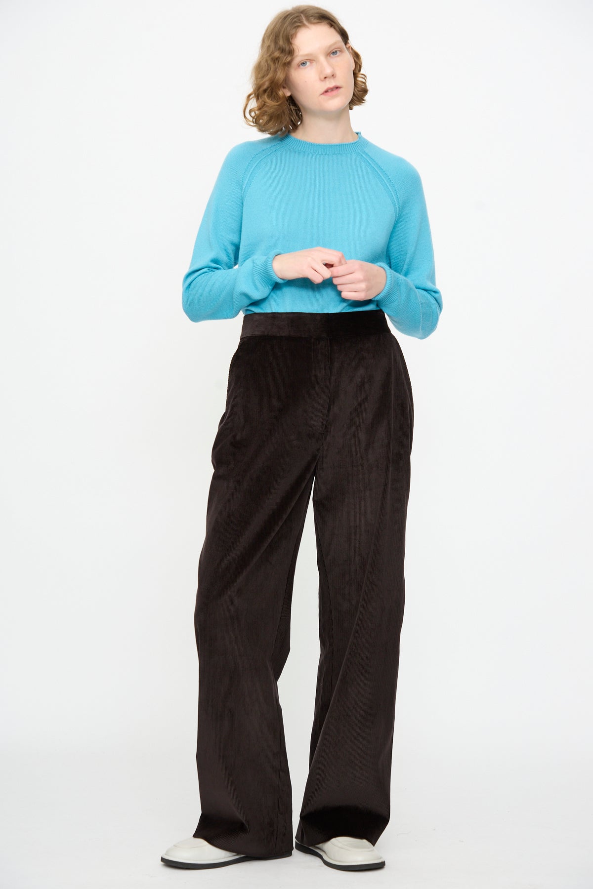 A person with short curly hair and a neutral expression stands against a white background, wearing a turquoise cotton sweater, Studio Nicholson’s Cotton Corduroy Grange Curved Pant in Espresso, and white shoes.