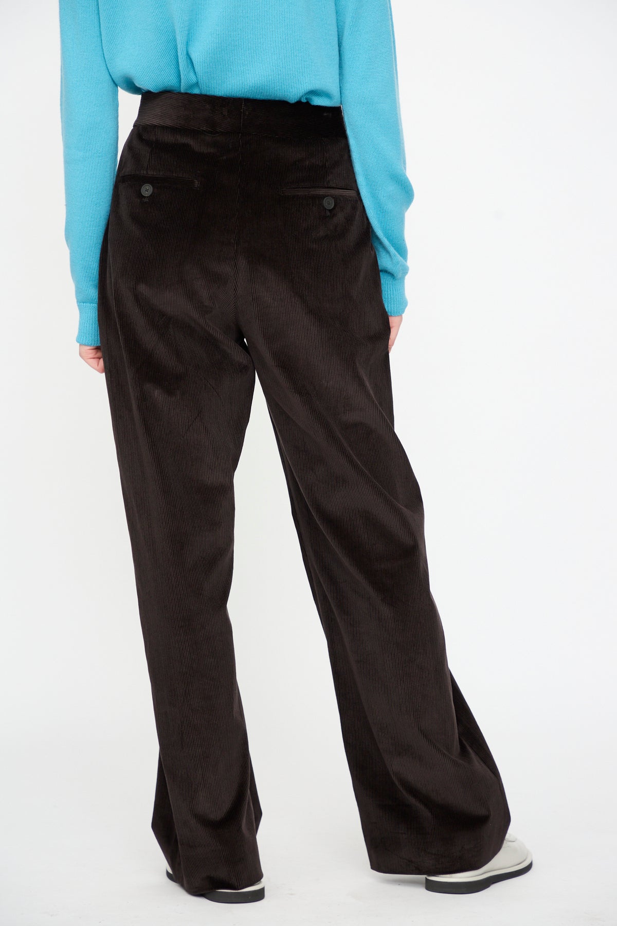 A person is shown from the back, wearing a blue long-sleeve top and Studio Nicholson’s Cotton Corduroy Grange Curved Pant in Espresso—a high-waisted, curved-leg style—standing with hands at their sides against a plain white background.