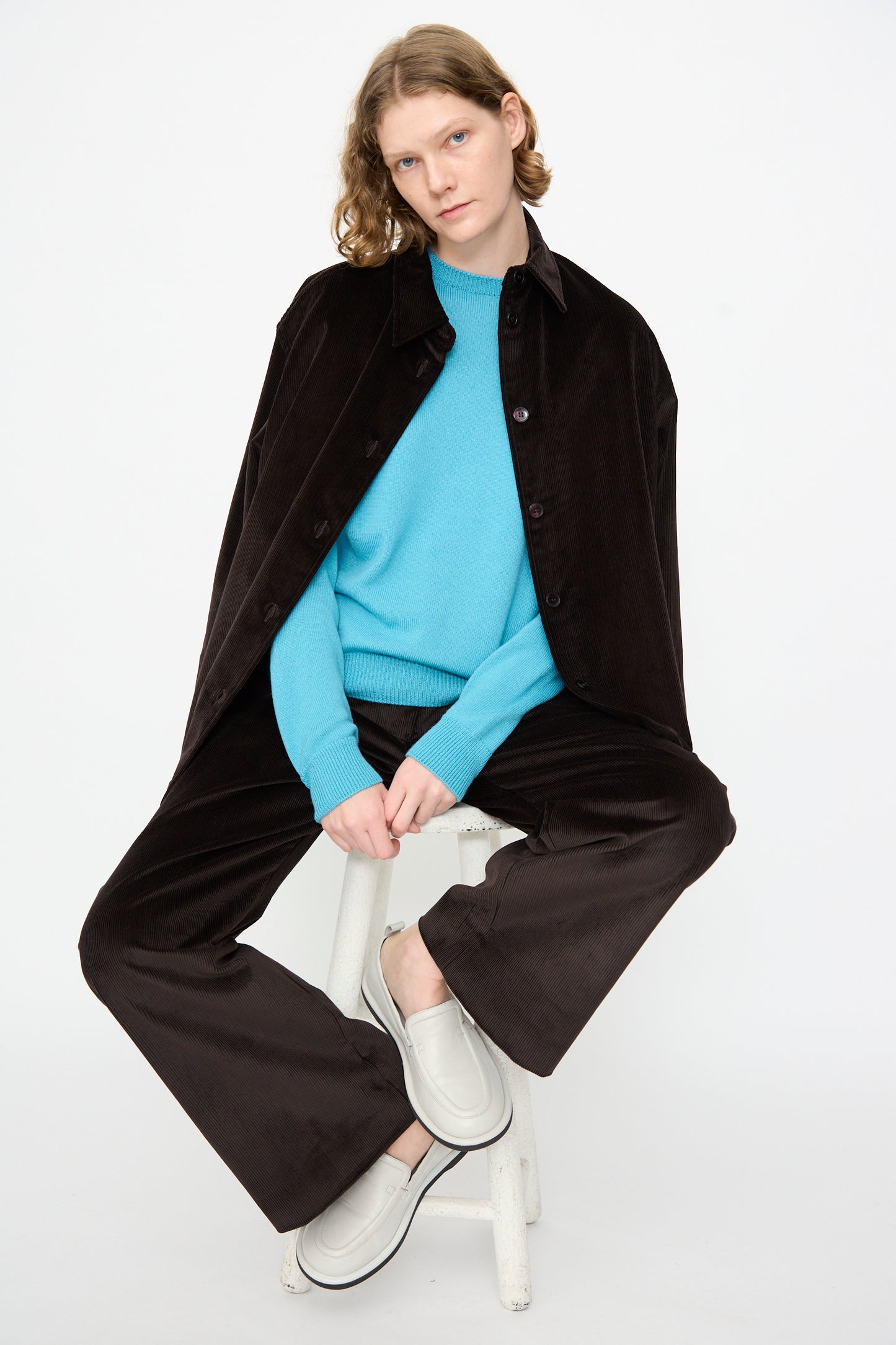 A person sits on a stool against a plain white background, wearing the Studio Nicholson Cotton Corduroy Santini Overshirt in Espresso with a relaxed fit, paired with matching black pants, a bright blue sweater, and white slip-on shoes.