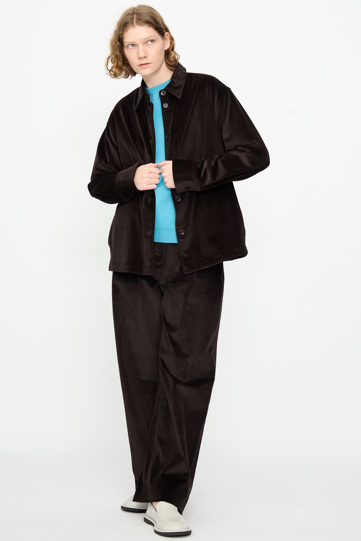 A person with long hair wears Studio Nicholson’s Cotton Corduroy Santini Overshirt in Espresso—a relaxed-fit black overshirt—paired with loose black pants, a turquoise shirt, and white slip-on shoes, standing against a plain white background. This look is made in Portugal.