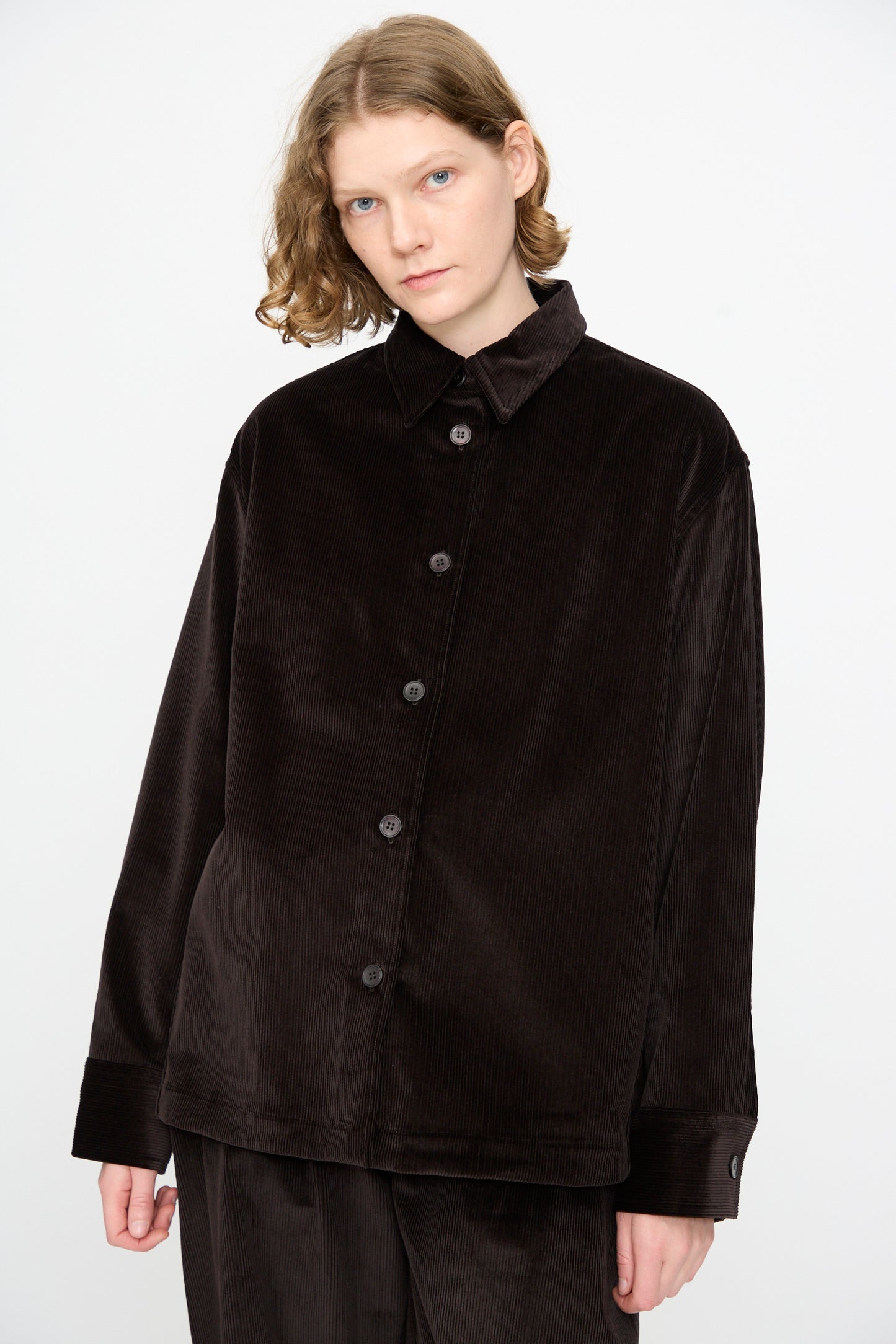 A person with short, wavy hair wears the Studio Nicholson Cotton Corduroy Santini Overshirt in Espresso—a relaxed-fit, buttoned, dark cotton corduroy overshirt—paired with matching pants, made in Portugal, standing against a plain white background.