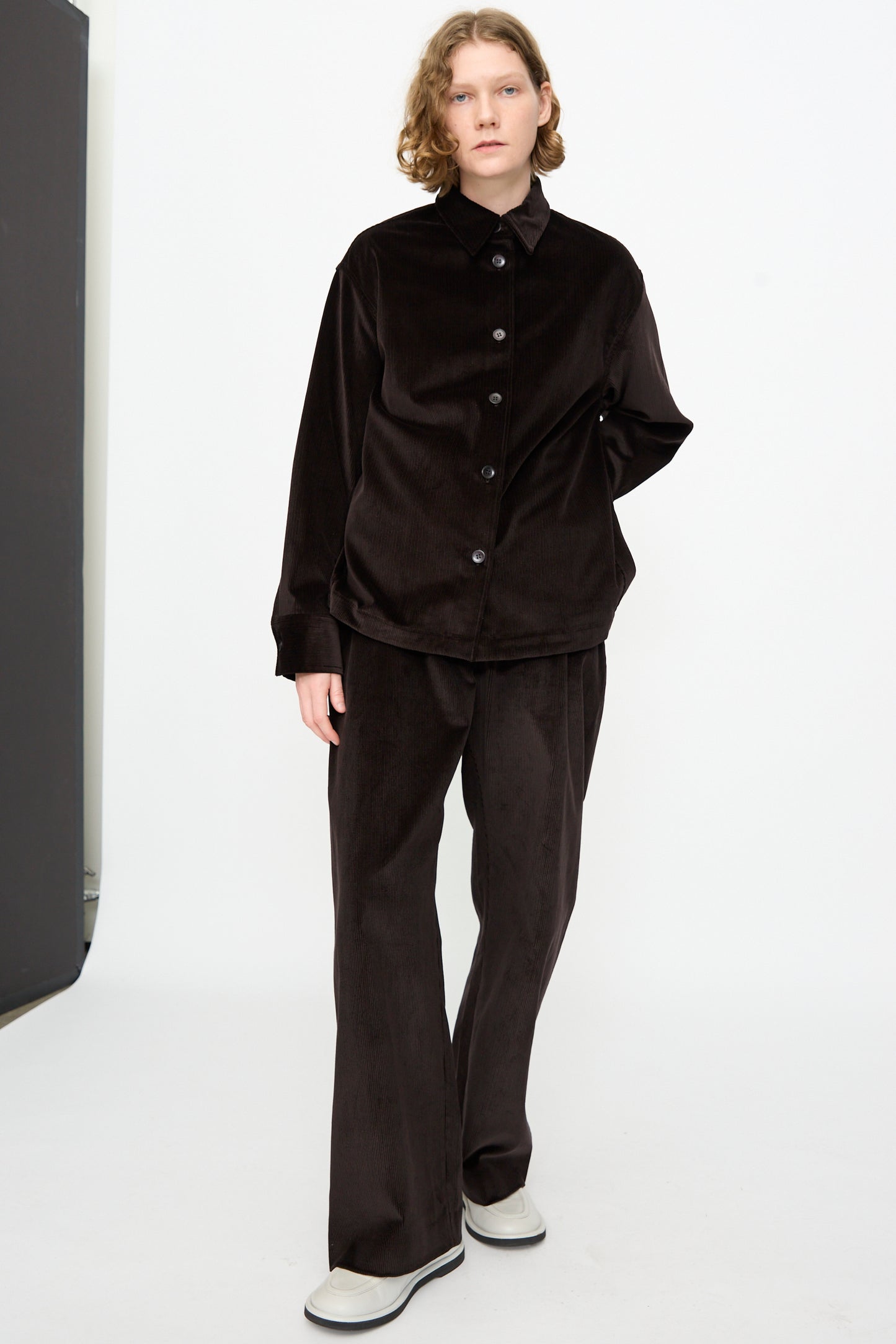 A person stands against a plain background wearing the Studio Nicholson Cotton Corduroy Santini Overshirt in Espresso, styled with matching pants and white shoes. The outfit features a relaxed fit and is made in Portugal.
