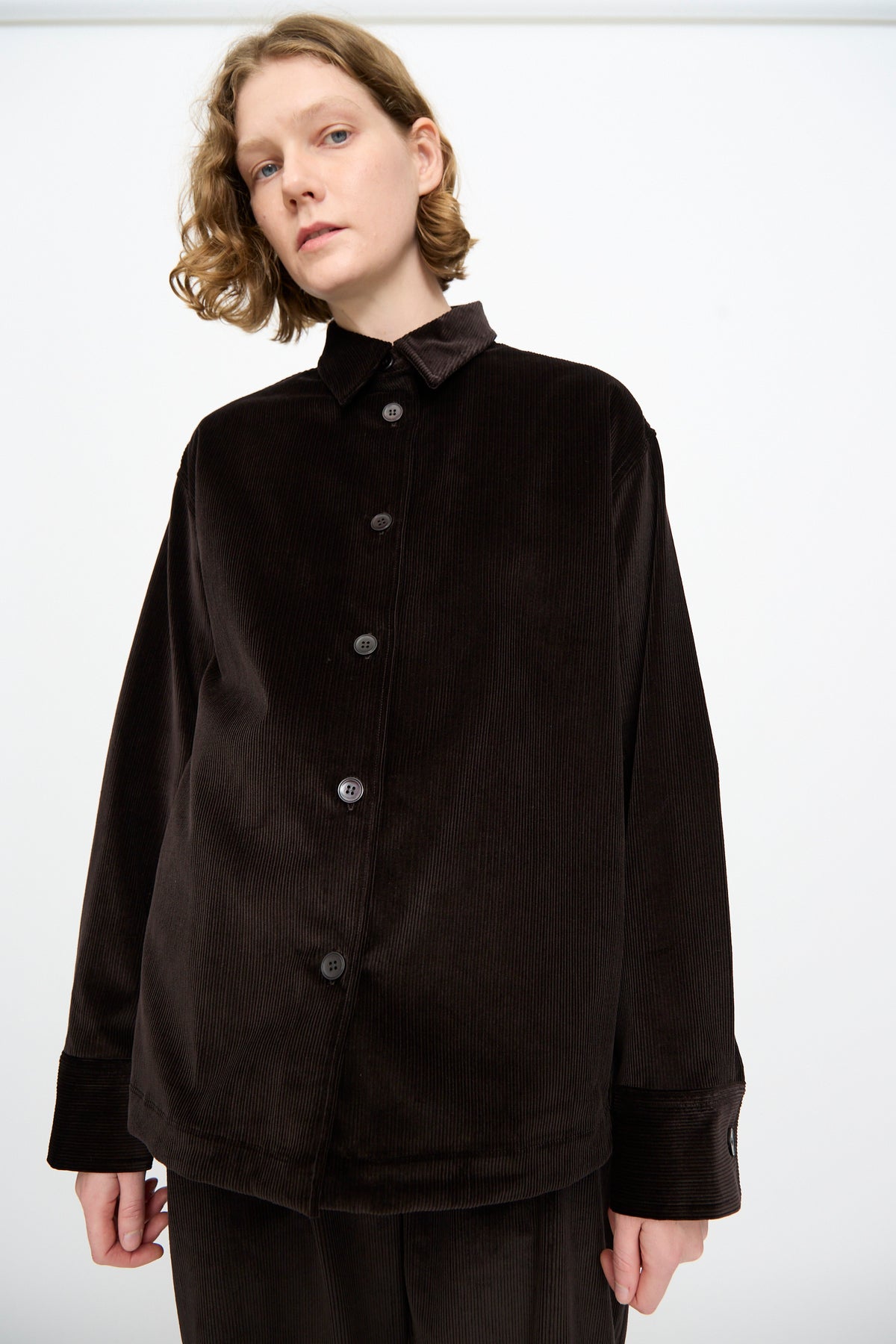 A person wearing the Studio Nicholson Cotton Corduroy Santini Overshirt in Espresso—a relaxed fit, long-sleeve design—stands against a plain white background.