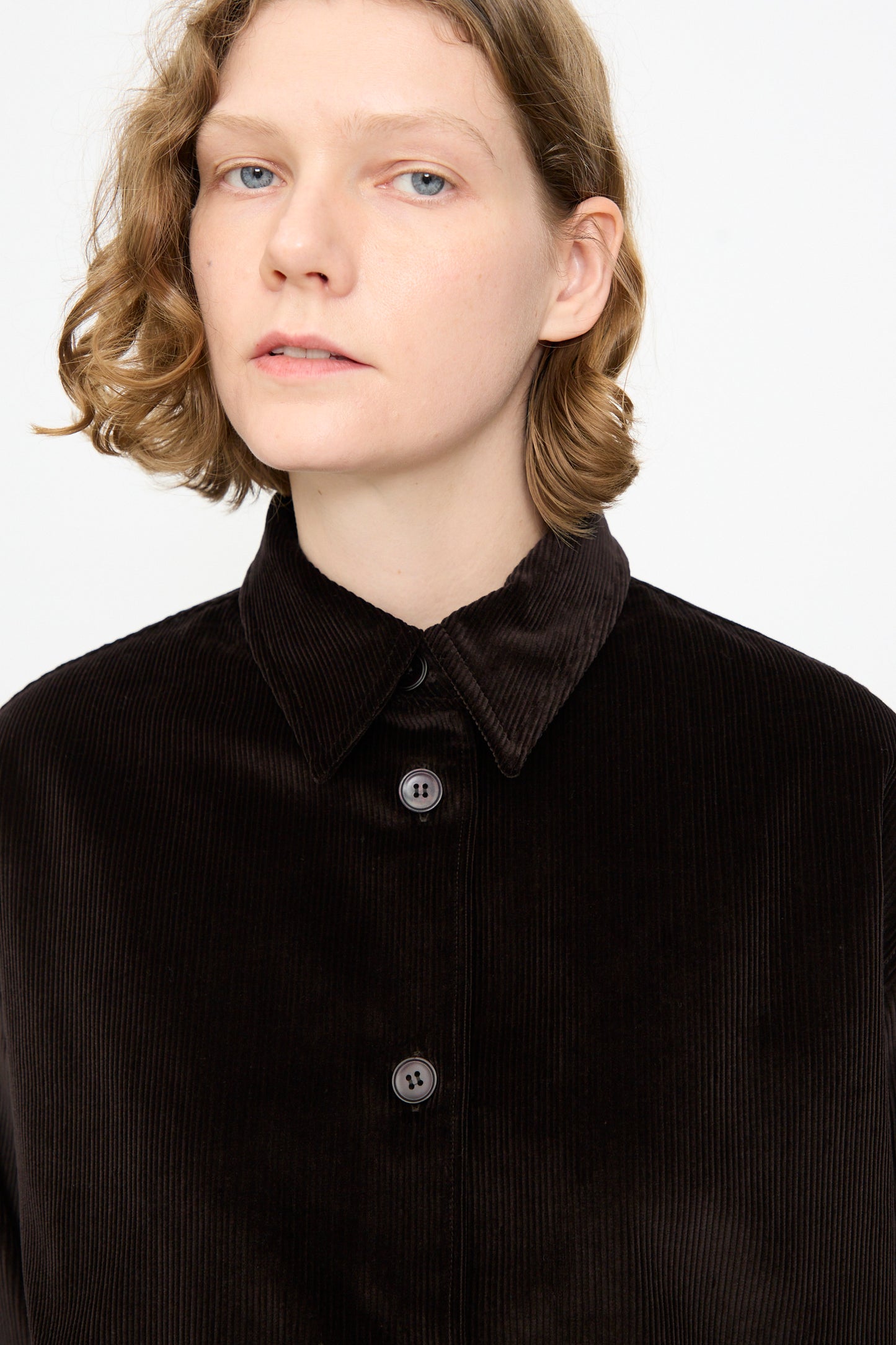 A person with short, wavy blond hair is wearing the Studio Nicholson Cotton Corduroy Santini Overshirt in Espresso, featuring a relaxed fit and crafted in Portugal; they are facing forward against a plain white background.