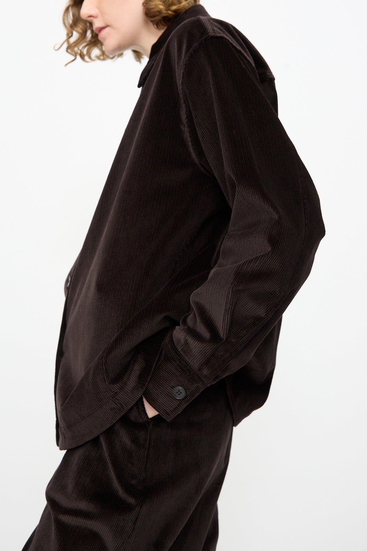 A person wearing the Studio Nicholson Cotton Corduroy Santini Overshirt in Espresso and matching trousers, photographed from the side with one hand in their pocket against a plain white background. The relaxed fit ensemble, crafted in Portugal, showcases effortless style and comfort.