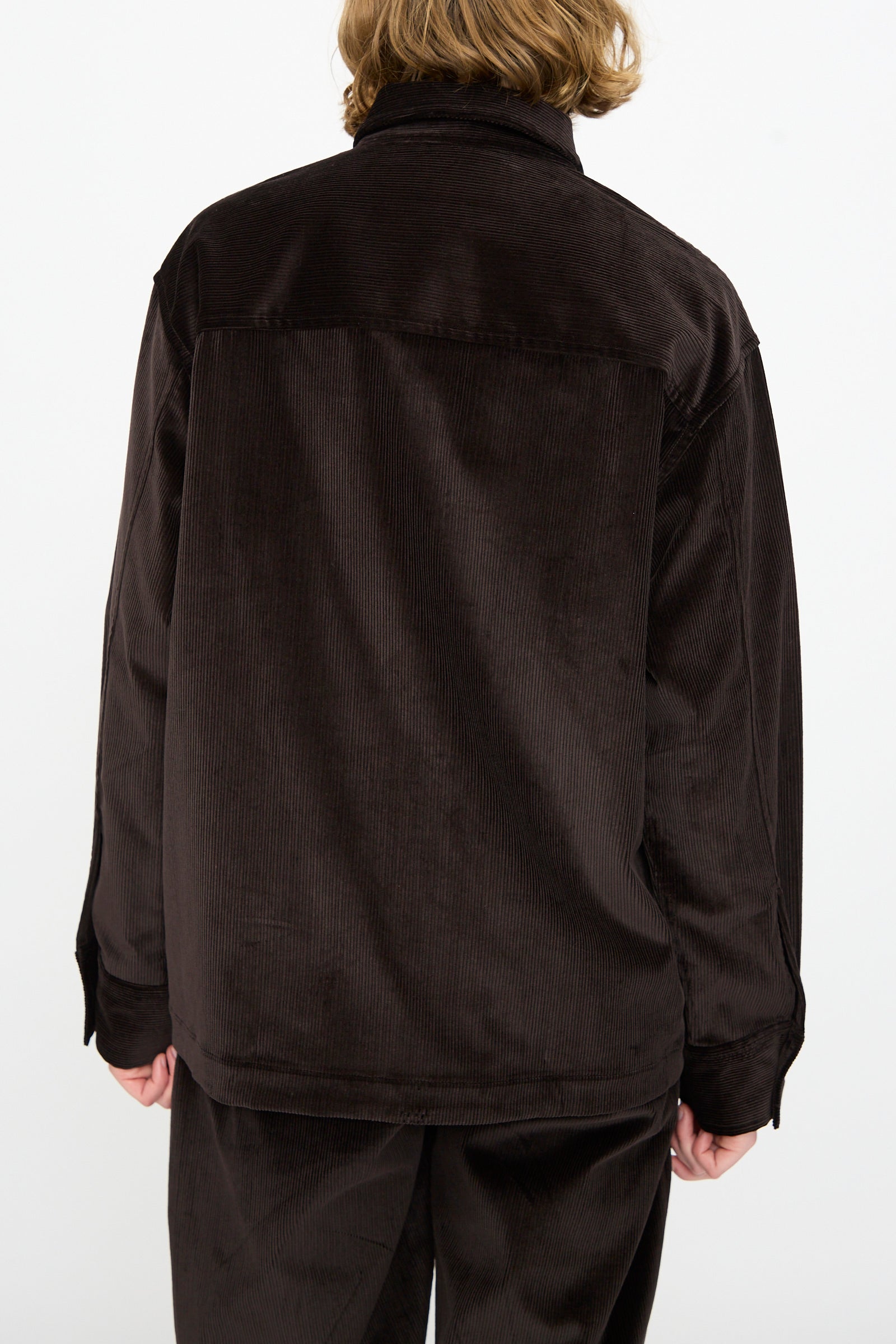A person with medium-length hair, shown from the back against a plain white background, is wearing the Studio Nicholson Cotton Corduroy Santini Overshirt in Espresso paired with matching pants in a relaxed fit.