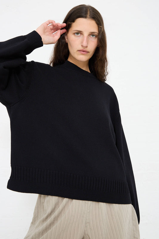 Person with long brown hair wearing a relaxed fit, Studio Nicholson Cotton Rib Paola Crewneck in Dark Navy and light beige pants poses with hand raised near head against a white background.