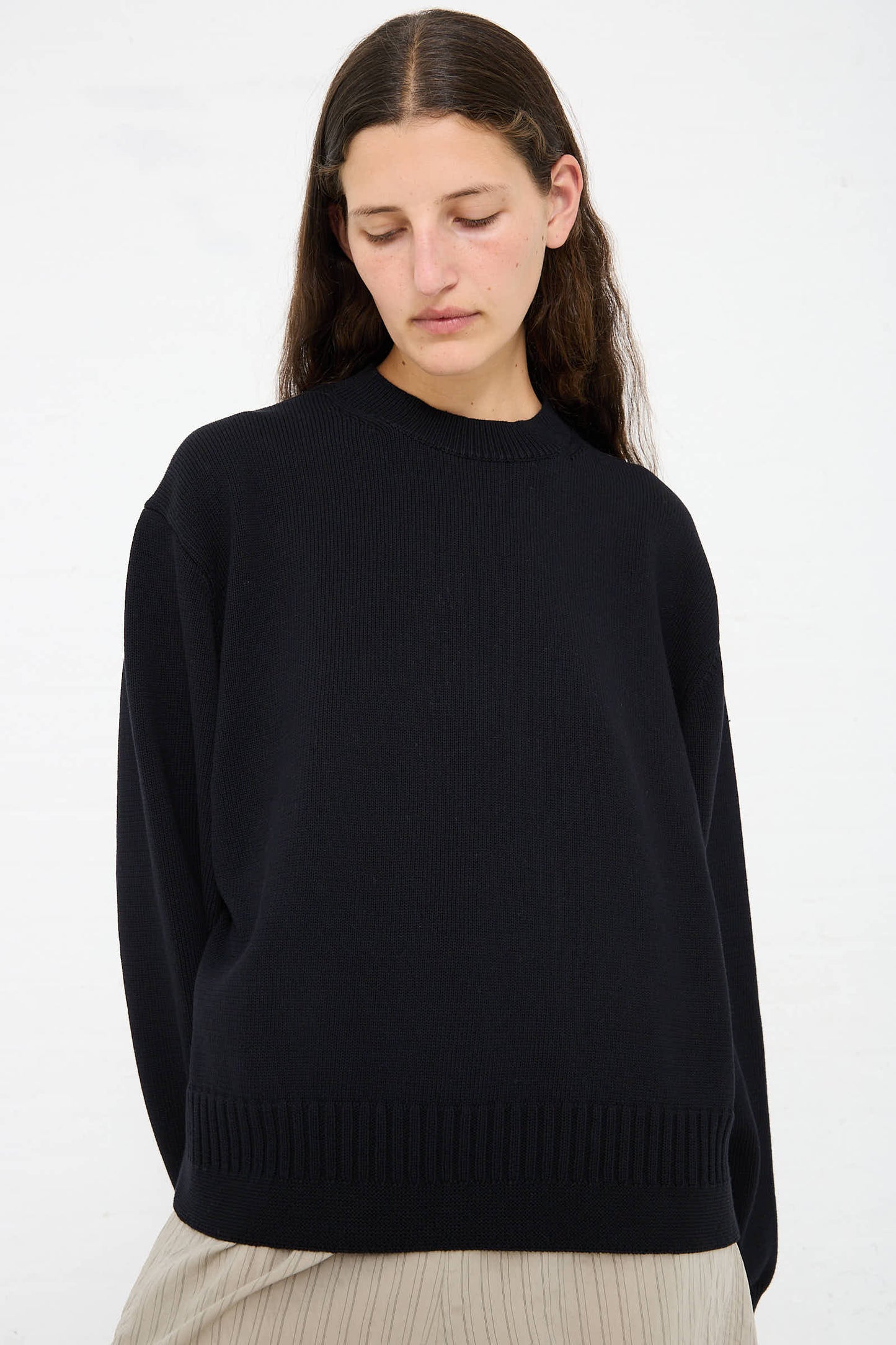 Person with long dark hair is wearing a relaxed fit, Cotton Rib Paola Crewneck in Dark Navy by Studio Nicholson and light-colored pants, looking downwards against a plain white background.