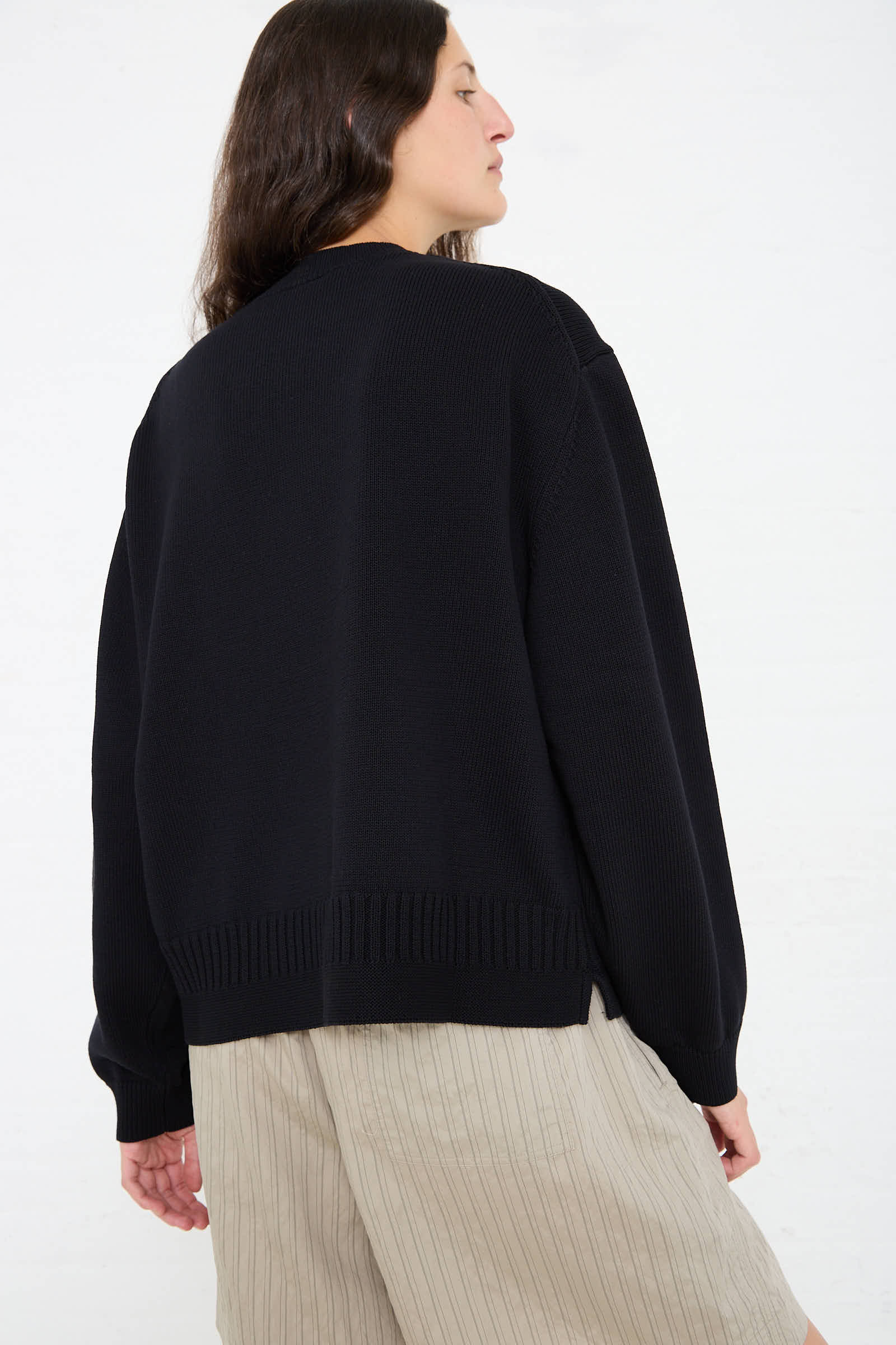 A person with long dark hair wearing a relaxed fit, Studio Nicholson Cotton Rib Paola Crewneck in Dark Navy and beige pants is seen from behind against a plain white background.