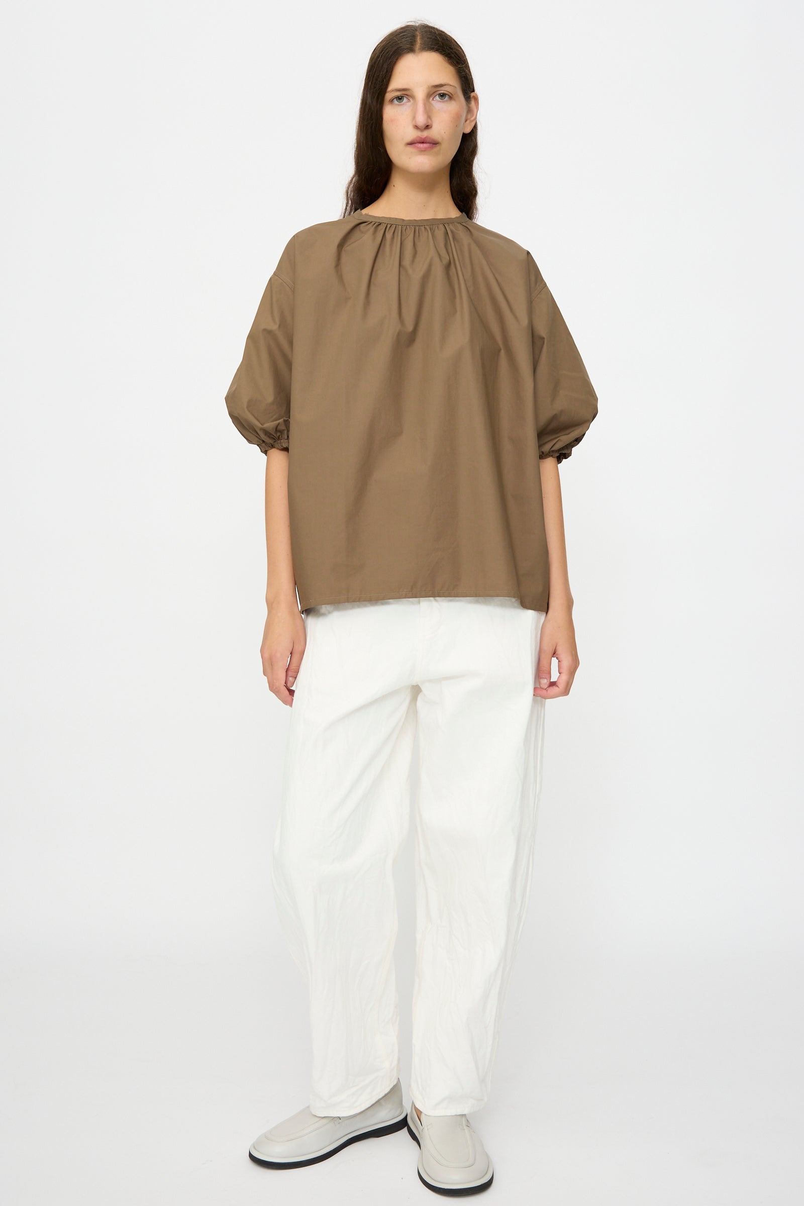 A person stands against a plain background wearing the Studio Nicholson Cove Gathered Top in Boulder, paired with white wide-leg pants and light-colored slip-on shoes.