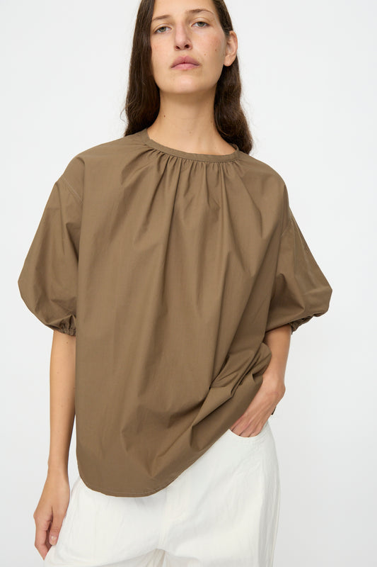 Wearing the Studio Nicholson Cove Gathered Top in Boulder, a person stands against a plain light background paired with white pants.