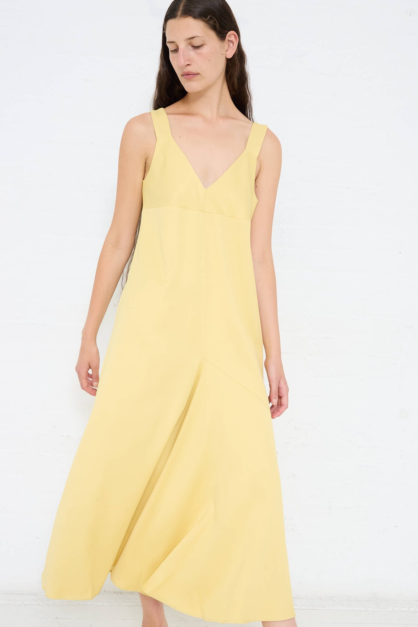 A person with long hair wearing a sleeveless, Crepe Wylie Asymmetric Slip Dress in Butter by Studio Nicholson with a deep V-neckline stands against a plain white background.