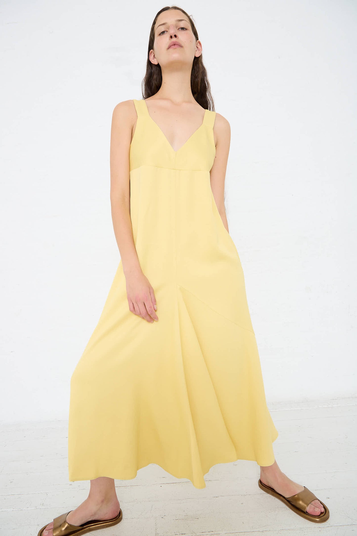 A person stands confidently against a white background, wearing a Crepe Wylie Asymmetric Slip Dress in Butter by Studio Nicholson and brown shoes.