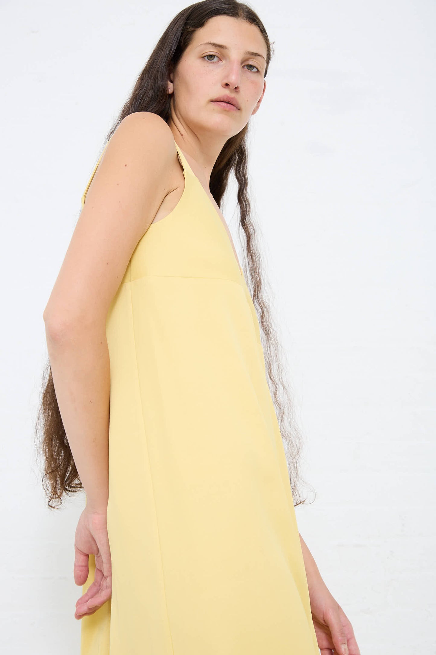 Person with long, dark hair wearing a sleeveless, Crepe Wylie Asymmetric Slip Dress in Butter from Studio Nicholson with a deep v-neckline, standing in front of a white background.