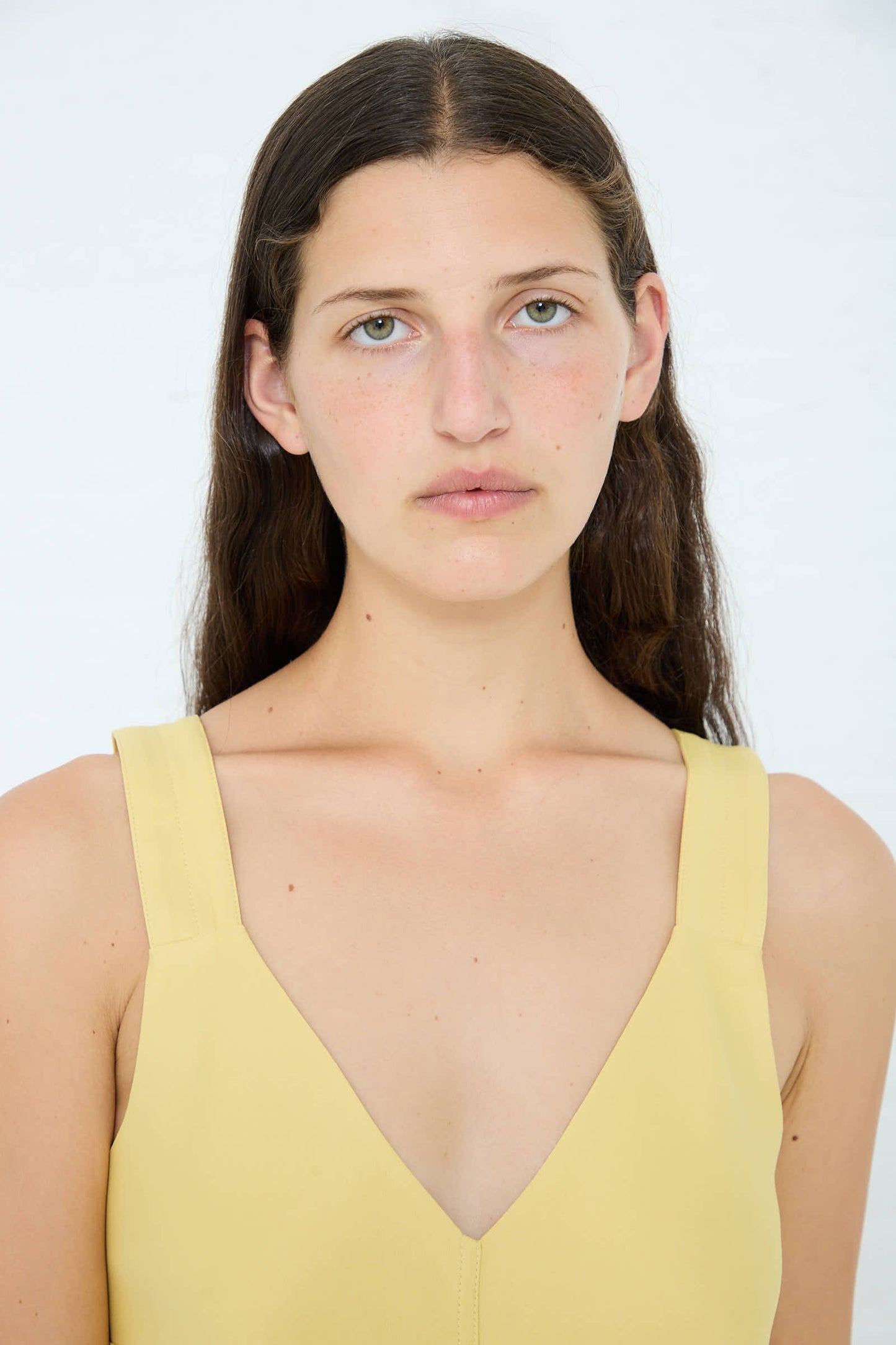 A woman with long dark hair and light eyes, wearing the Studio Nicholson Crepe Wylie Asymmetric Slip Dress in Butter, looks directly at the camera against a plain white background.