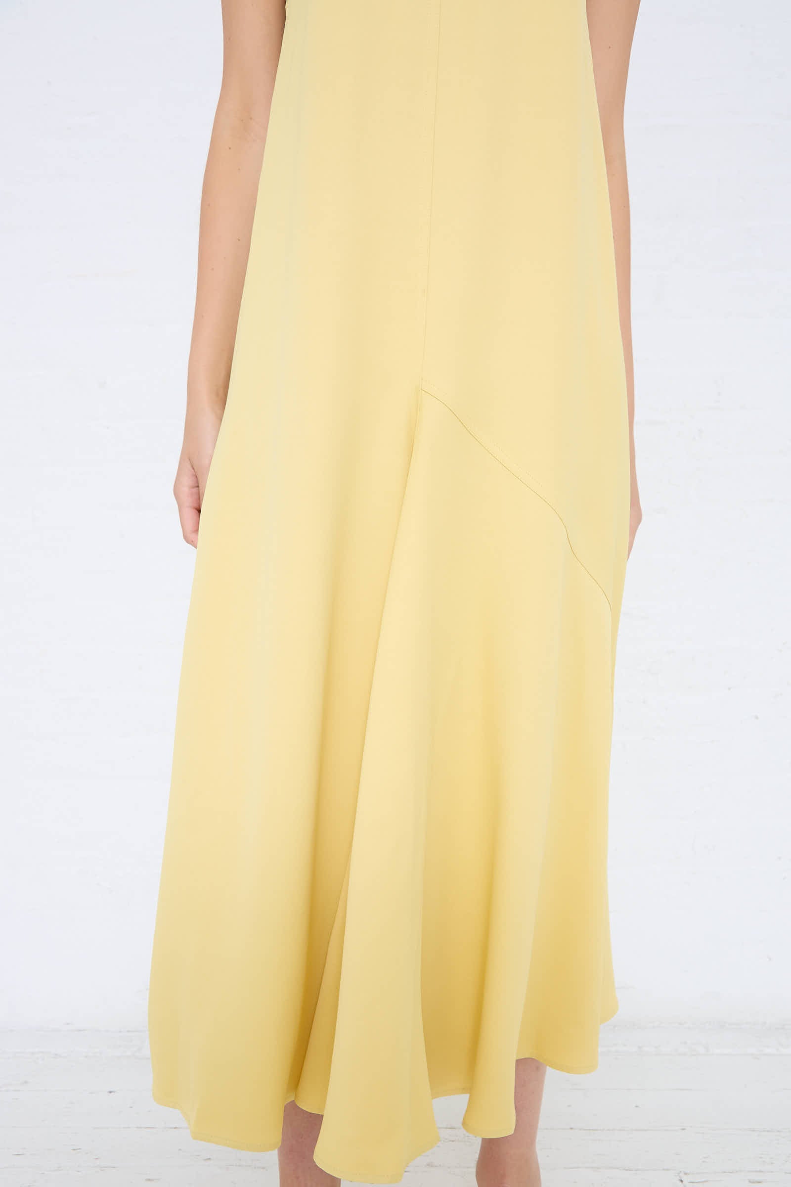 A person wearing Studio Nicholson's Crepe Wylie Asymmetric Slip Dress in Butter made of viscose crepe with a flowing skirt and deep V-neckline, standing against a plain white background.