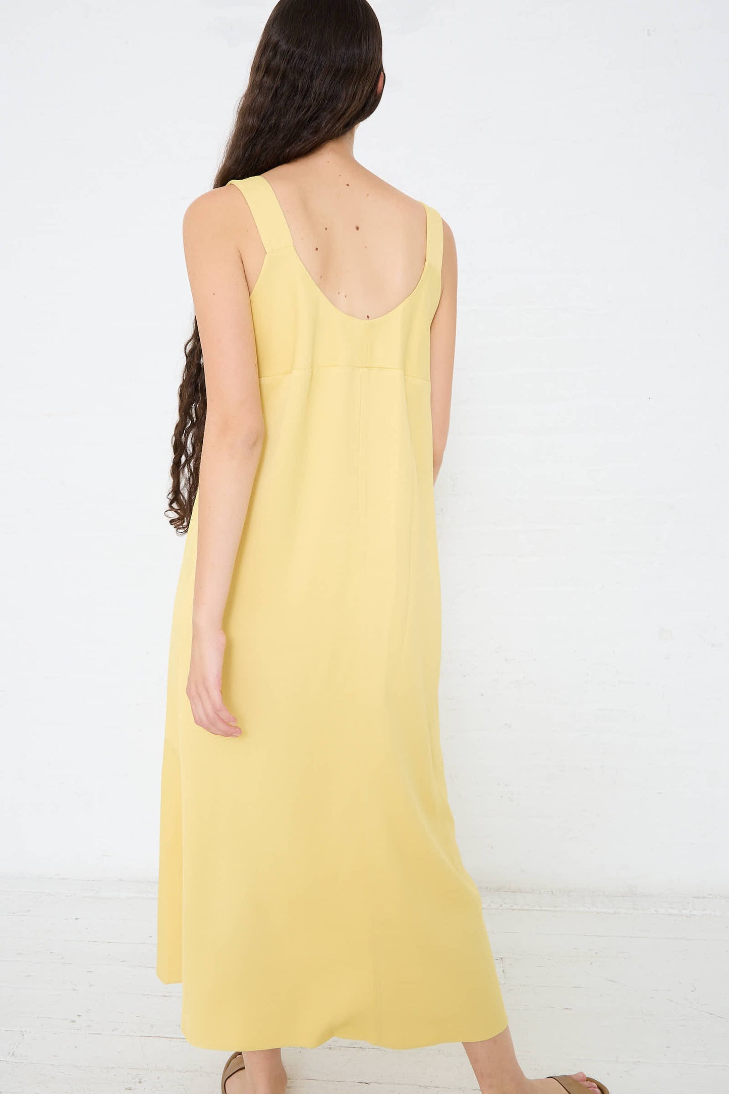 A woman with long hair wearing a sleeveless yellow Crepe Wylie Asymmetric Slip Dress in Butter by Studio Nicholson and sandals is standing with her back to the camera against a plain white background.