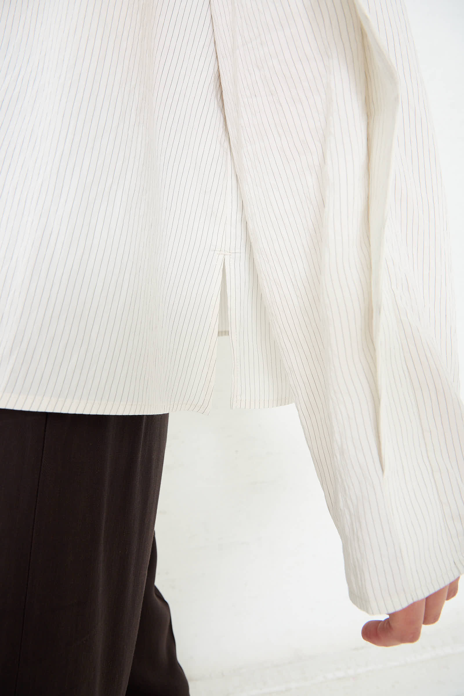 A close-up of a person wearing the Studio Nicholson Emmet Drape Sleeve Top in Black and White Stripe and dark pants, showing part of the draped sleeve and shirt hem.