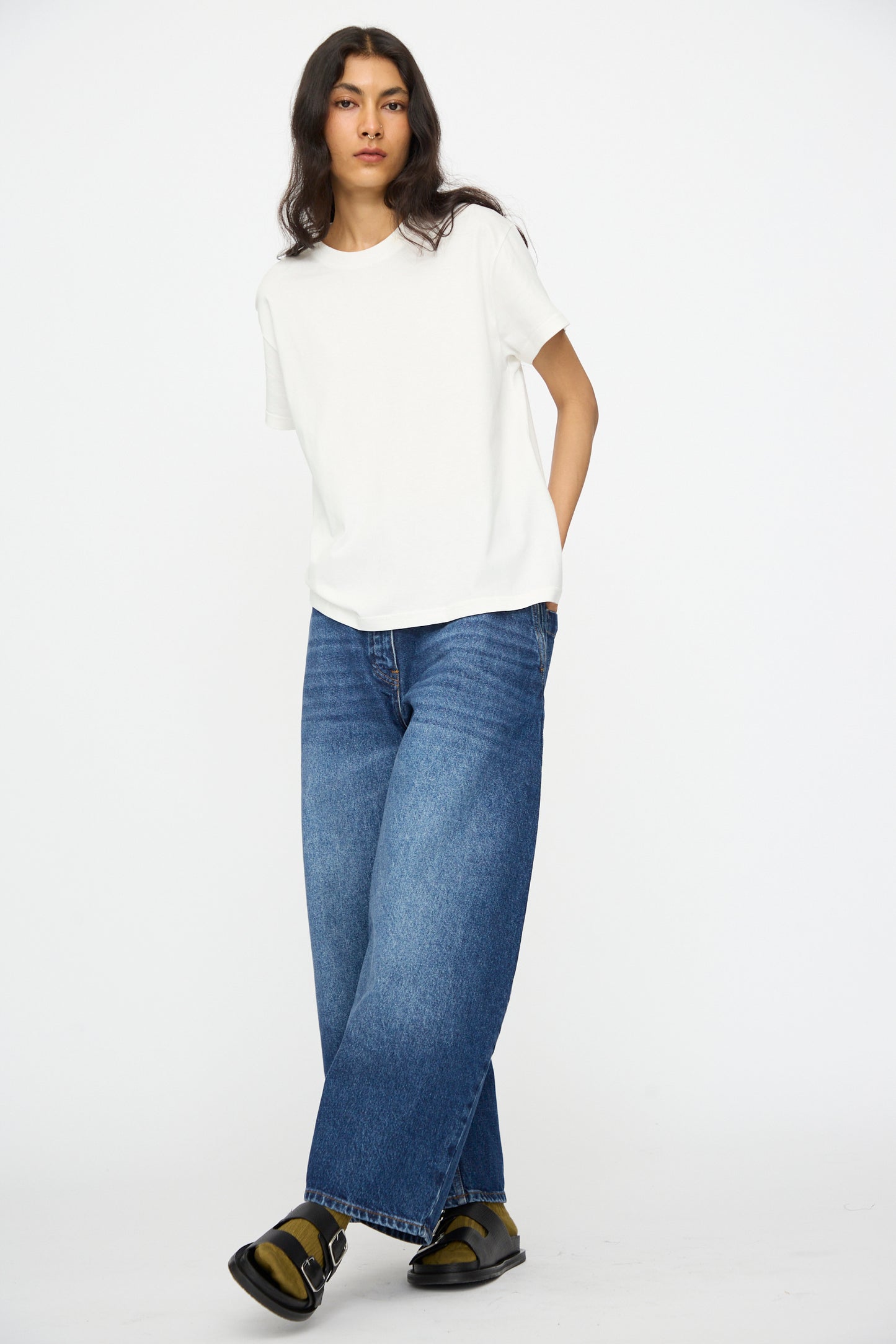 A person stands against a plain white background, wearing a white t-shirt, Studio Nicholson Denim Chalco Jean in Worn Wash Indigo, and black sandals.