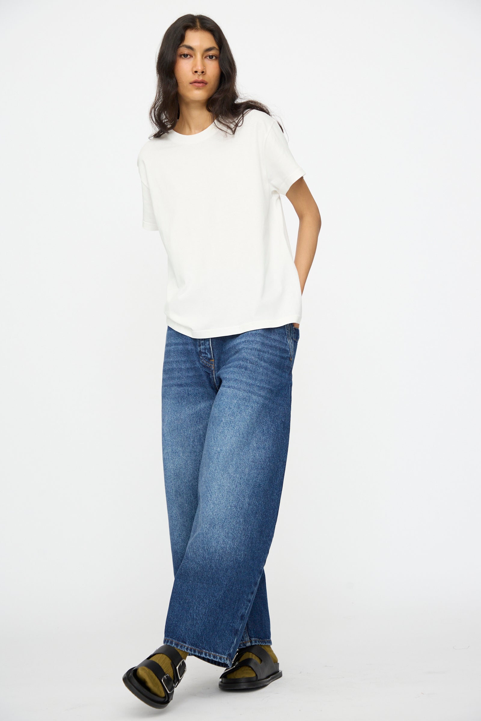 A person stands against a plain white background, wearing a white t-shirt, Studio Nicholson Denim Chalco Jean in Worn Wash Indigo, and black sandals.