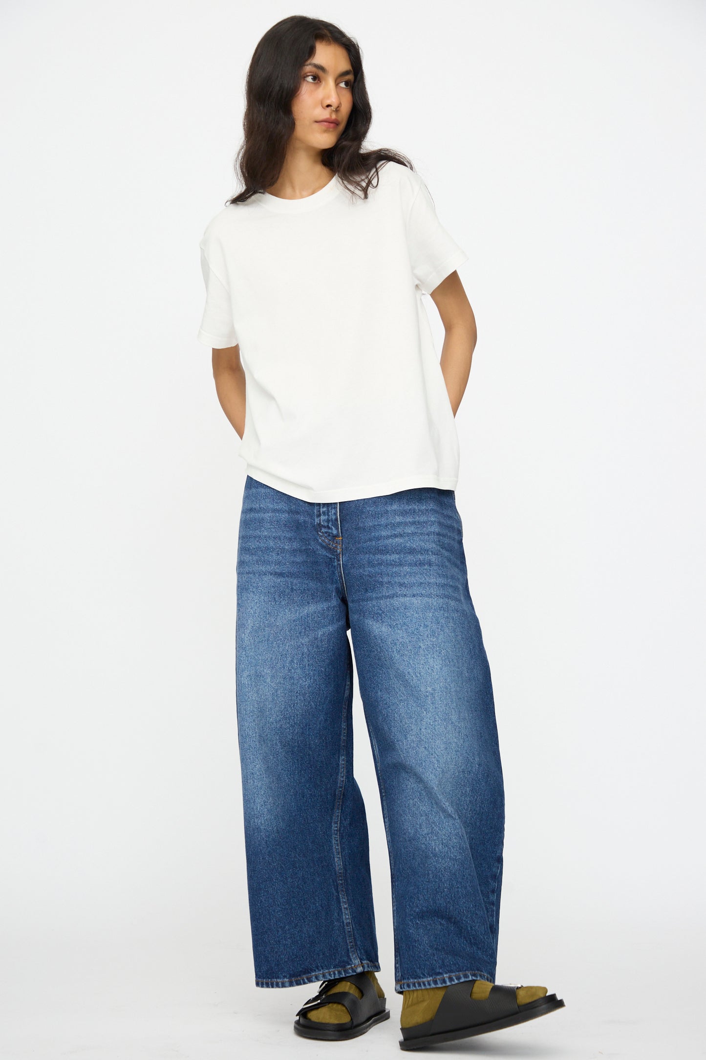 A person wearing a plain white t-shirt, Studio Nicholson's Denim Chalco Jean in Worn Wash Indigo with an oversized fit, and sandals stands against a plain white background.