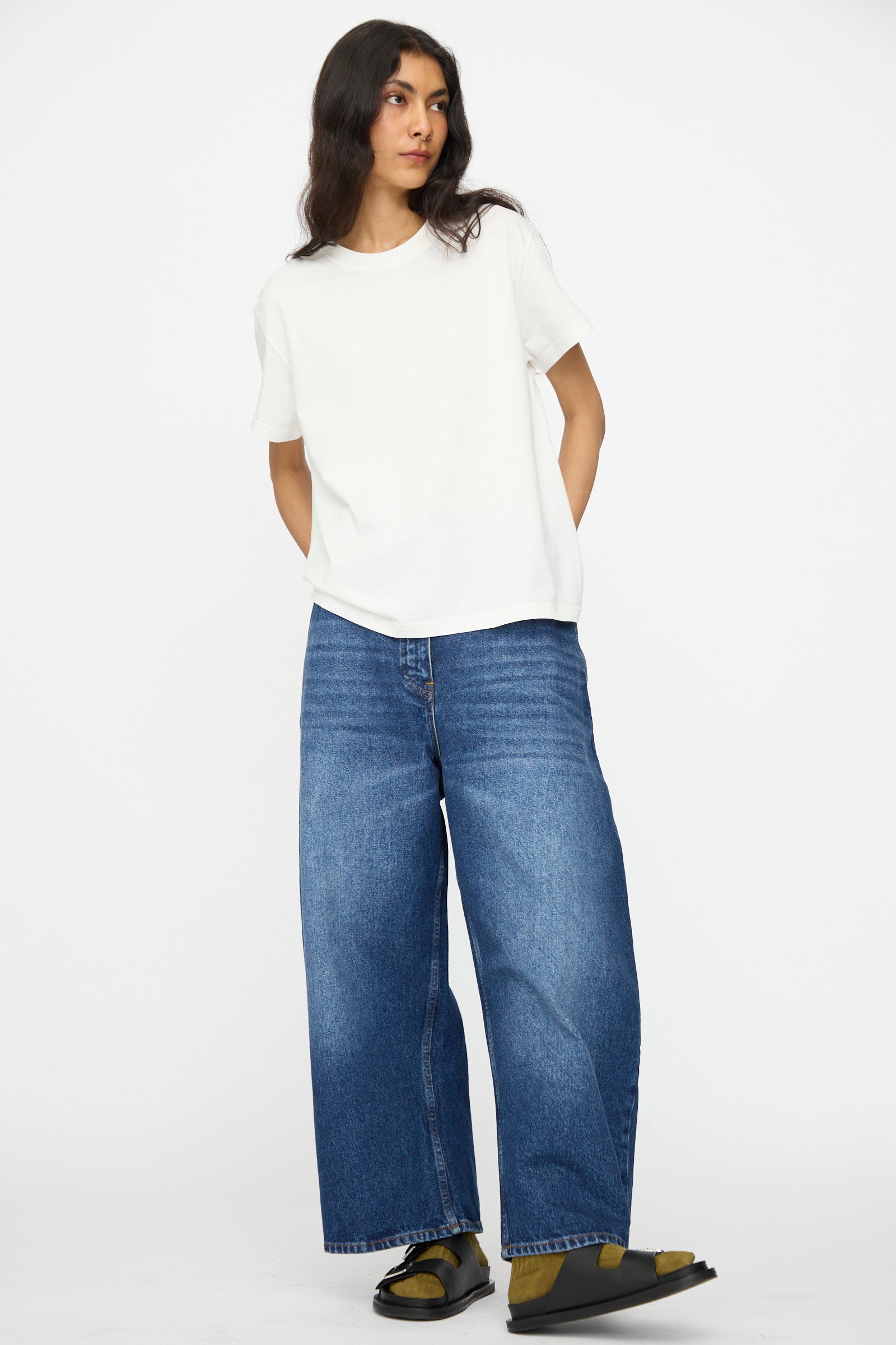 A person wearing a plain white t-shirt, Studio Nicholson's Denim Chalco Jean in Worn Wash Indigo with an oversized fit, and sandals stands against a plain white background.