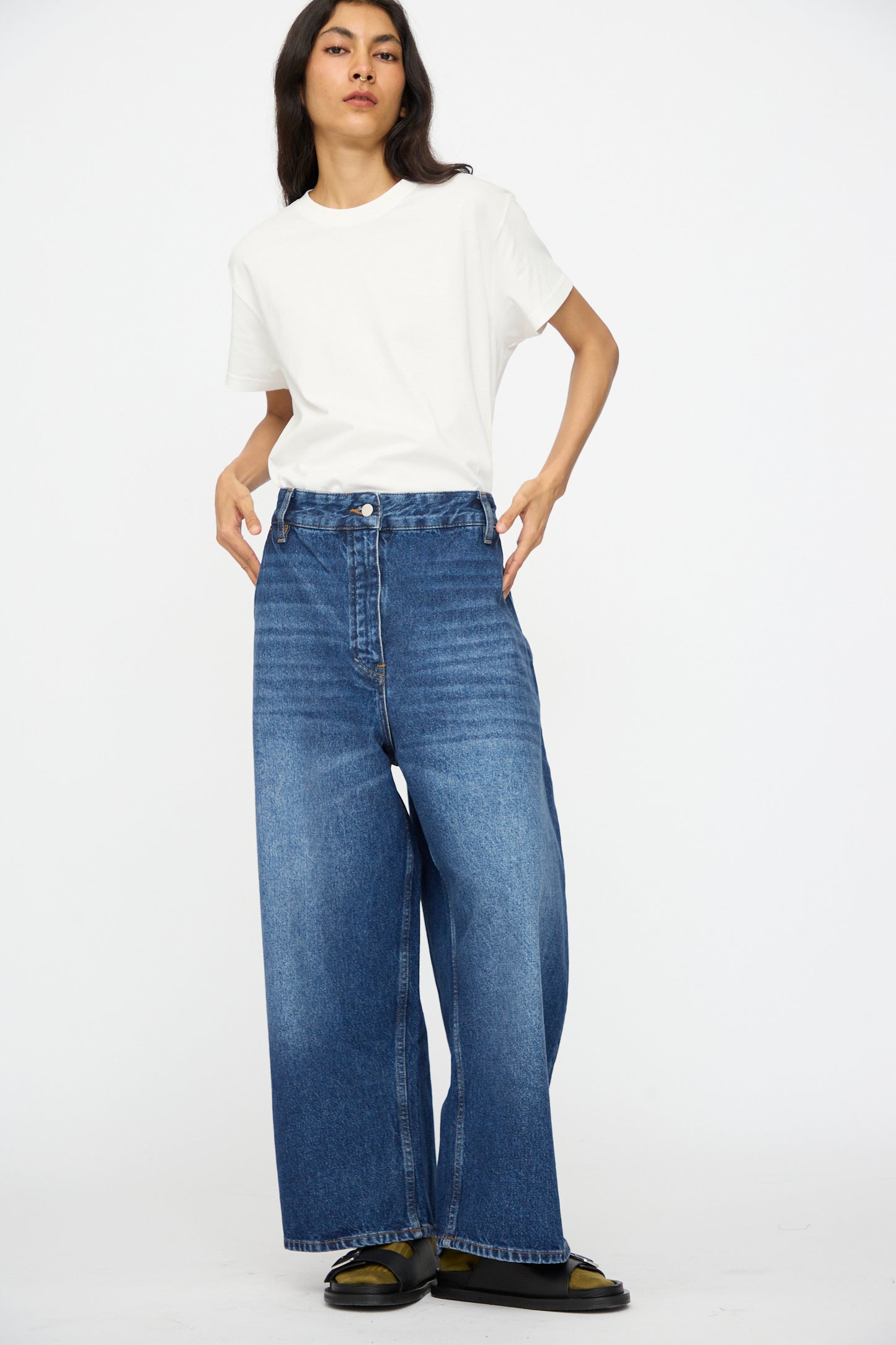 A person wearing a plain white t-shirt, Studio Nicholson Denim Chalco Jean in Worn Wash Indigo, and black shoes stands against a plain white background.