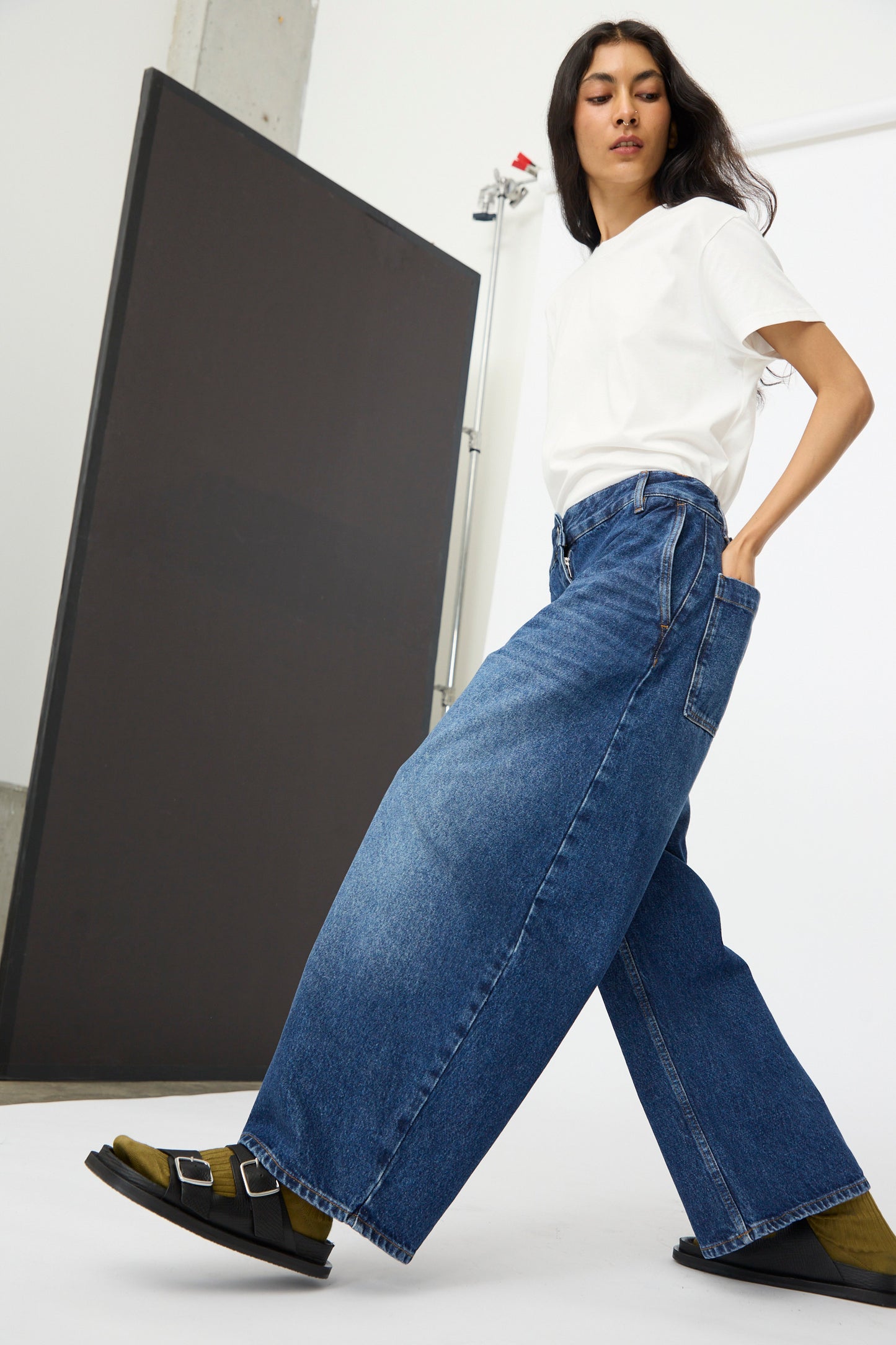 A woman stands in a minimalist studio wearing a white t-shirt, Studio Nicholson's Denim Chalco Jean in Worn Wash Indigo, green socks, and black sandals.