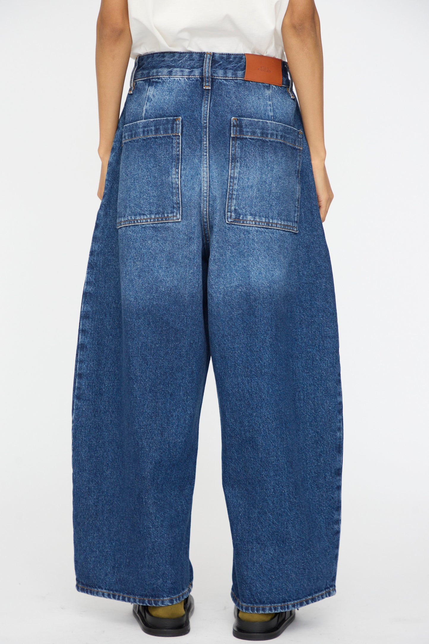 Model shown from behind wearing Studio Nicholson’s Denim Chalco Jean in Worn Wash Indigo, an oversized, wide leg style with large back pockets and leather patch at the waistband, paired with black shoes.