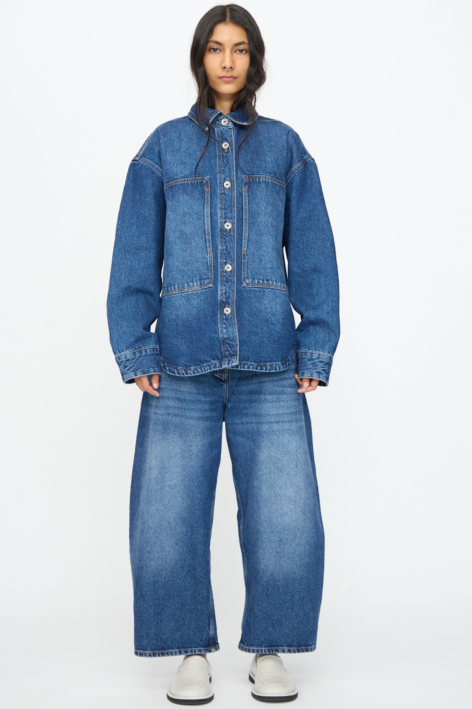 A person stands facing forward in the Studio Nicholson Denim Paso Shirt in Worn Wash Indigo and wide-leg denim jeans with white shoes, against a plain white background.