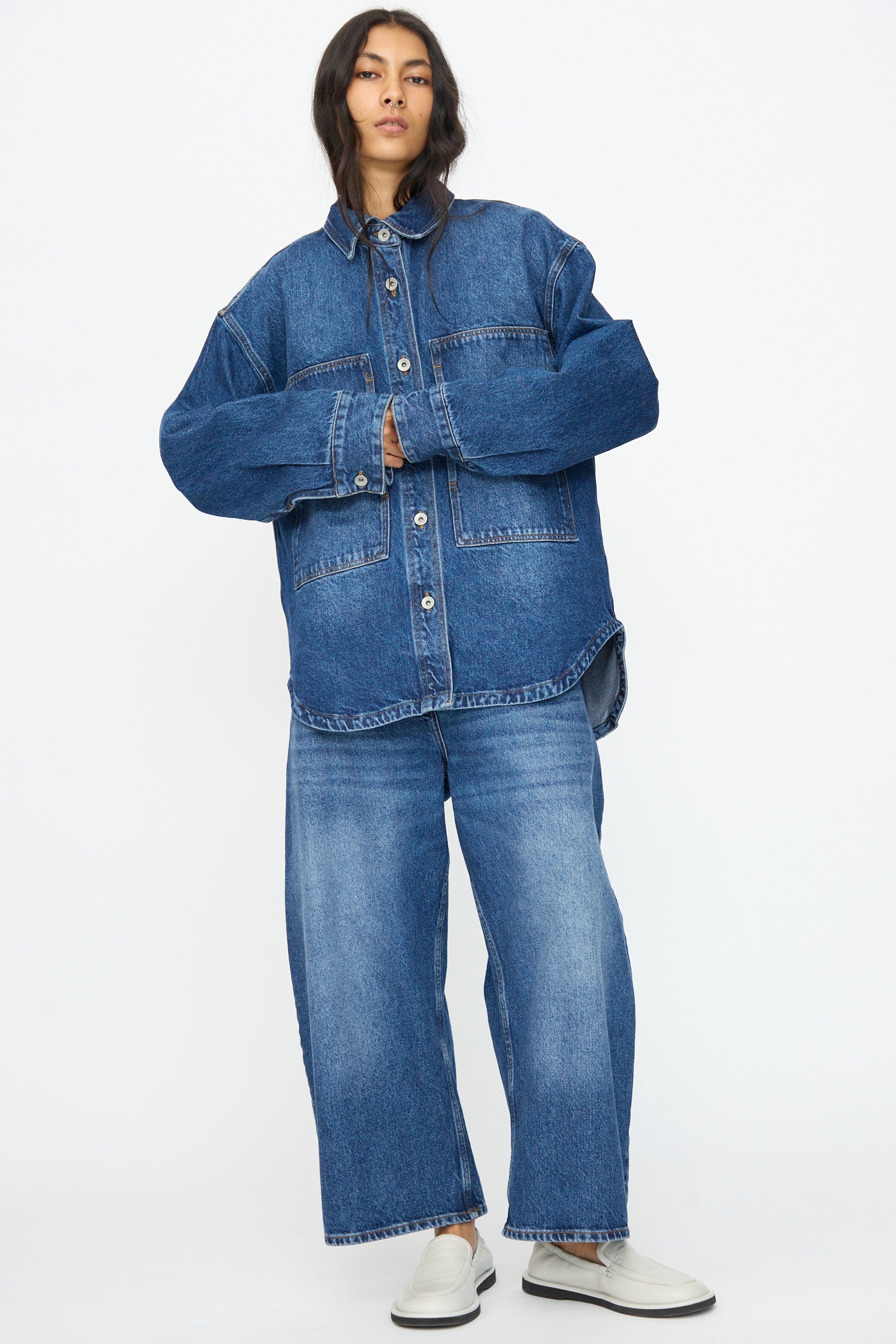 A person stands against a plain background wearing the Studio Nicholson Denim Paso Shirt in Worn Wash Indigo, paired with matching wide-leg jeans and white slip-on shoes.