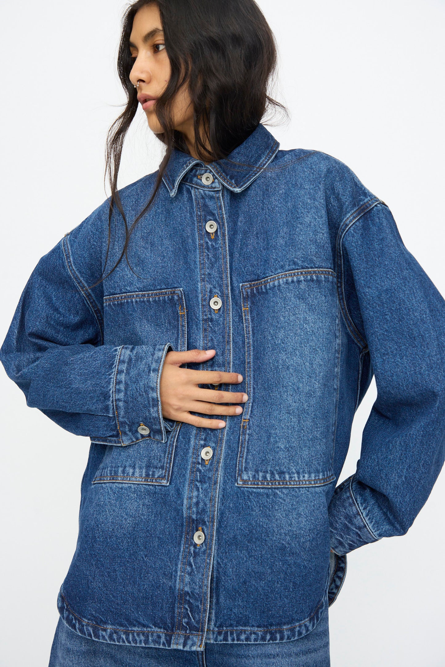 A person wears the Studio Nicholson Denim Paso Shirt in Worn Wash Indigo, featuring oversized patch pockets, standing against a plain white background.