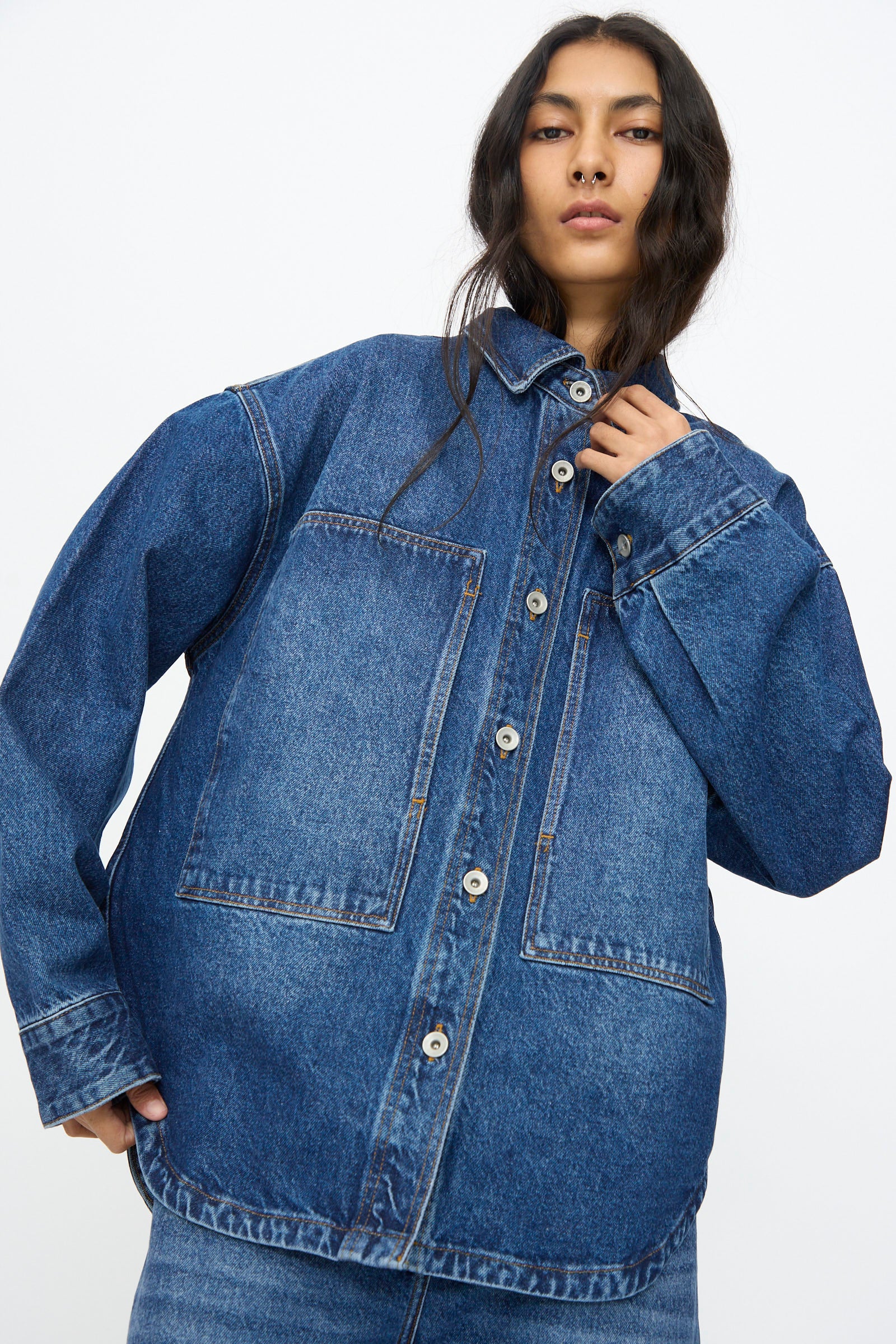 A person wears the Studio Nicholson Denim Paso Shirt in Worn Wash Indigo, featuring oversized patch pockets, paired with jeans and standing against a plain white background.