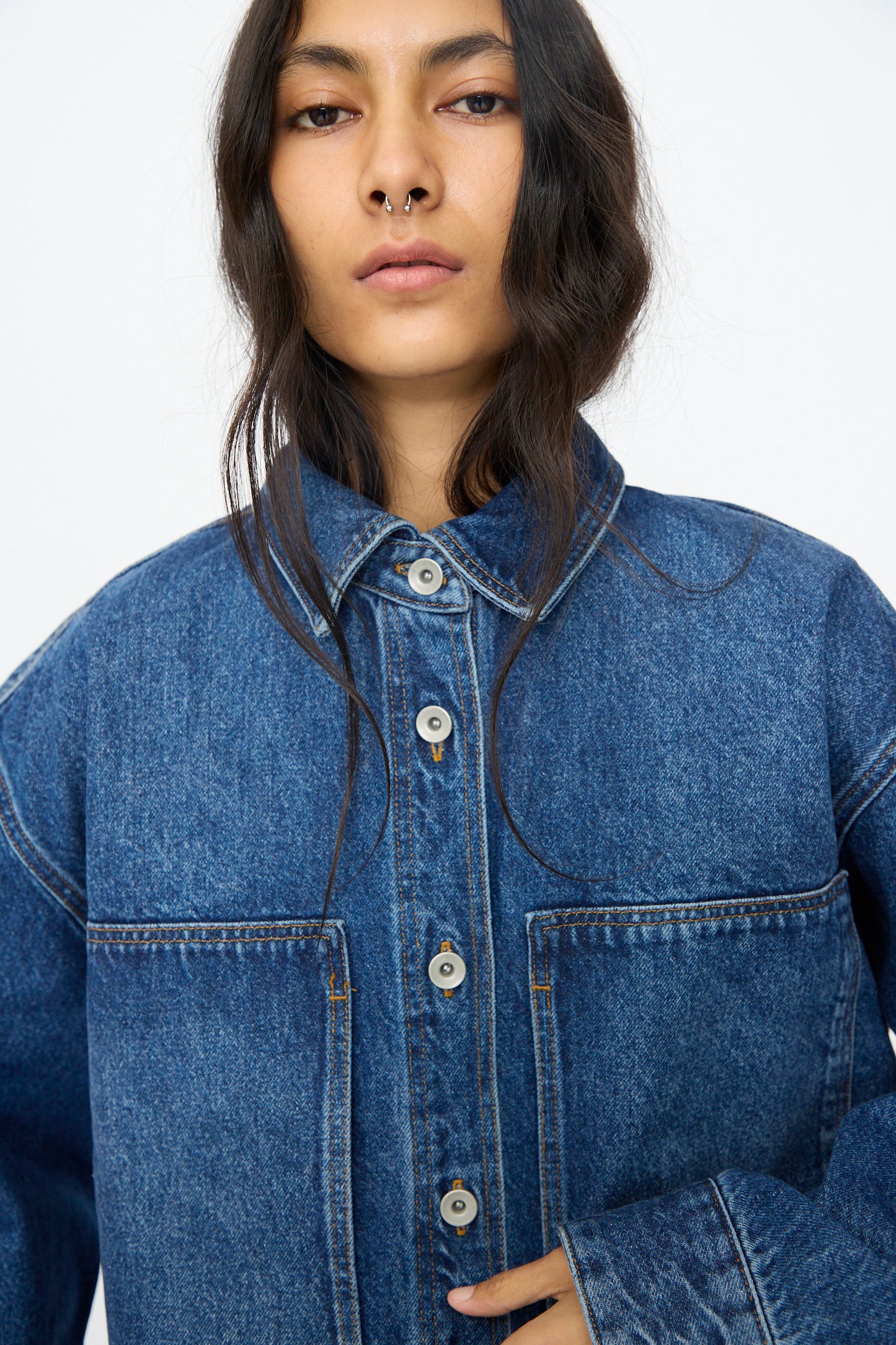 Person with long dark hair and a neutral expression, wearing the Studio Nicholson Denim Paso Shirt in Worn Wash Indigo with oversized patch pockets, looking directly at the camera.