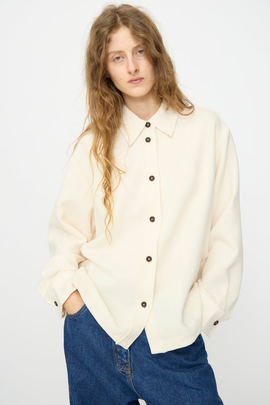 A person with long wavy hair wears the Studio Nicholson Double Wool Sablé Santini Overshirt in Chalk and blue jeans, standing against a plain light background.