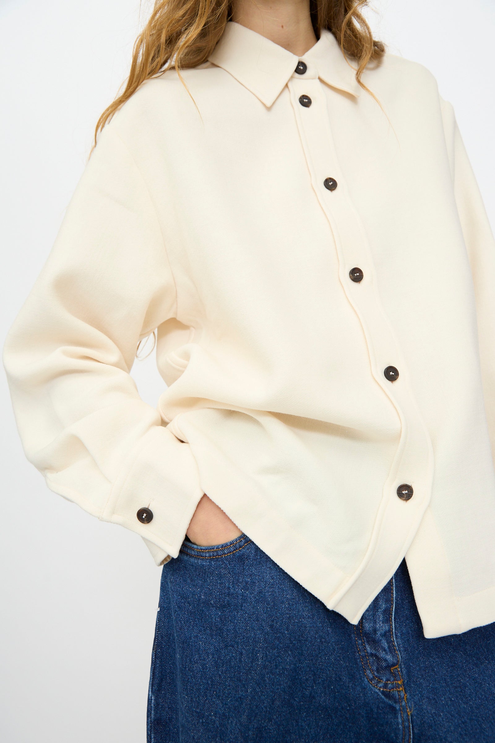 Wearing the Studio Nicholson Double Wool Sablé Santini Overshirt in Chalk with dark buttons and blue jeans, this person poses with one hand in their pocket against a plain background.