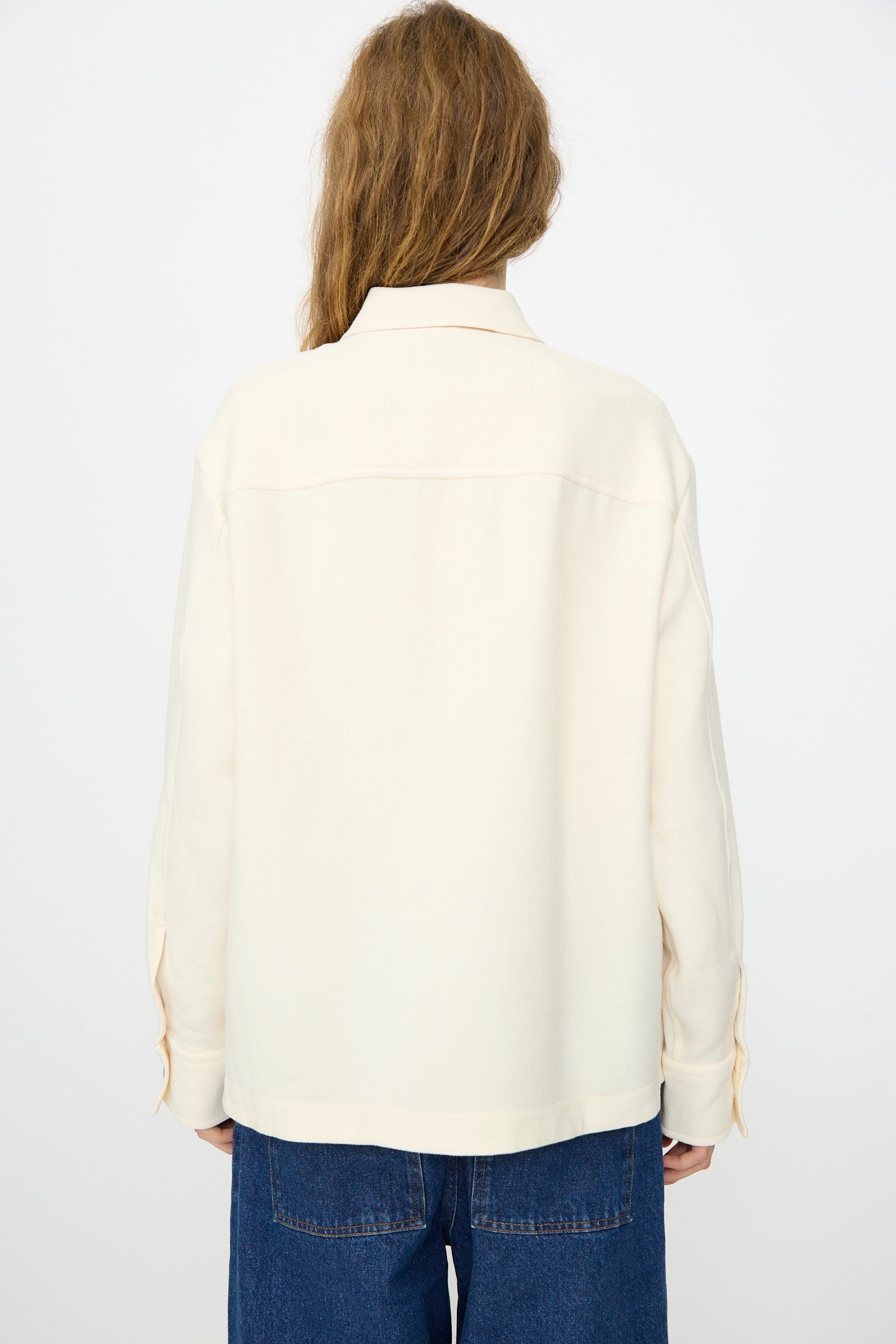 A person with long hair, seen from behind, wears the Studio Nicholson Double Wool Sablé Santini Overshirt in Chalk and blue jeans against a plain light background.